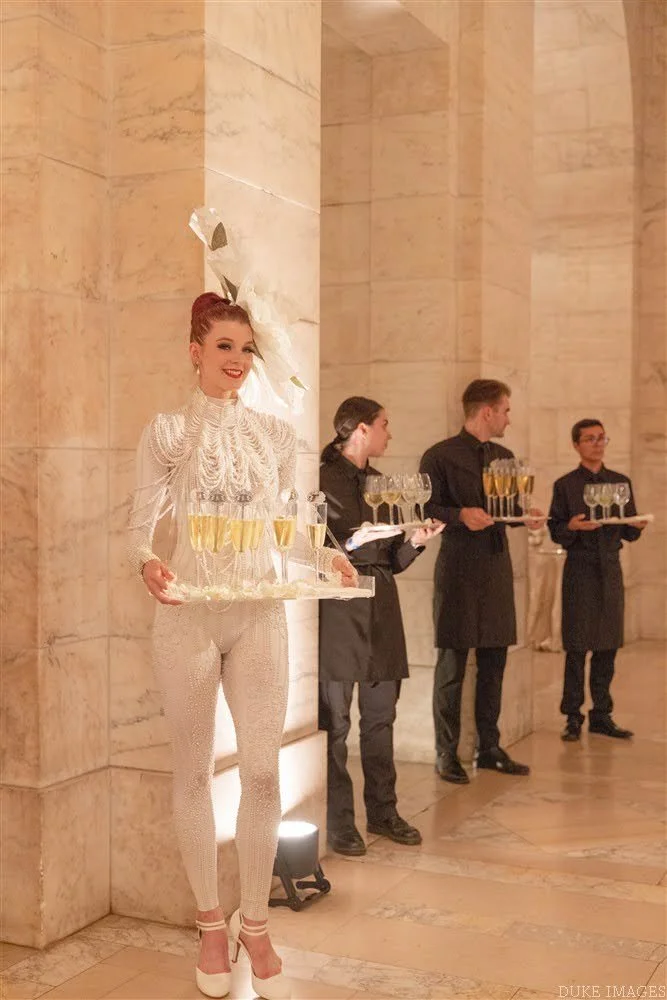 A woman in an ornate white outfit holding a tray with glasses of champagne at a formal event, standing next to three waitstaff who are also holding trays with glasses of champagne.