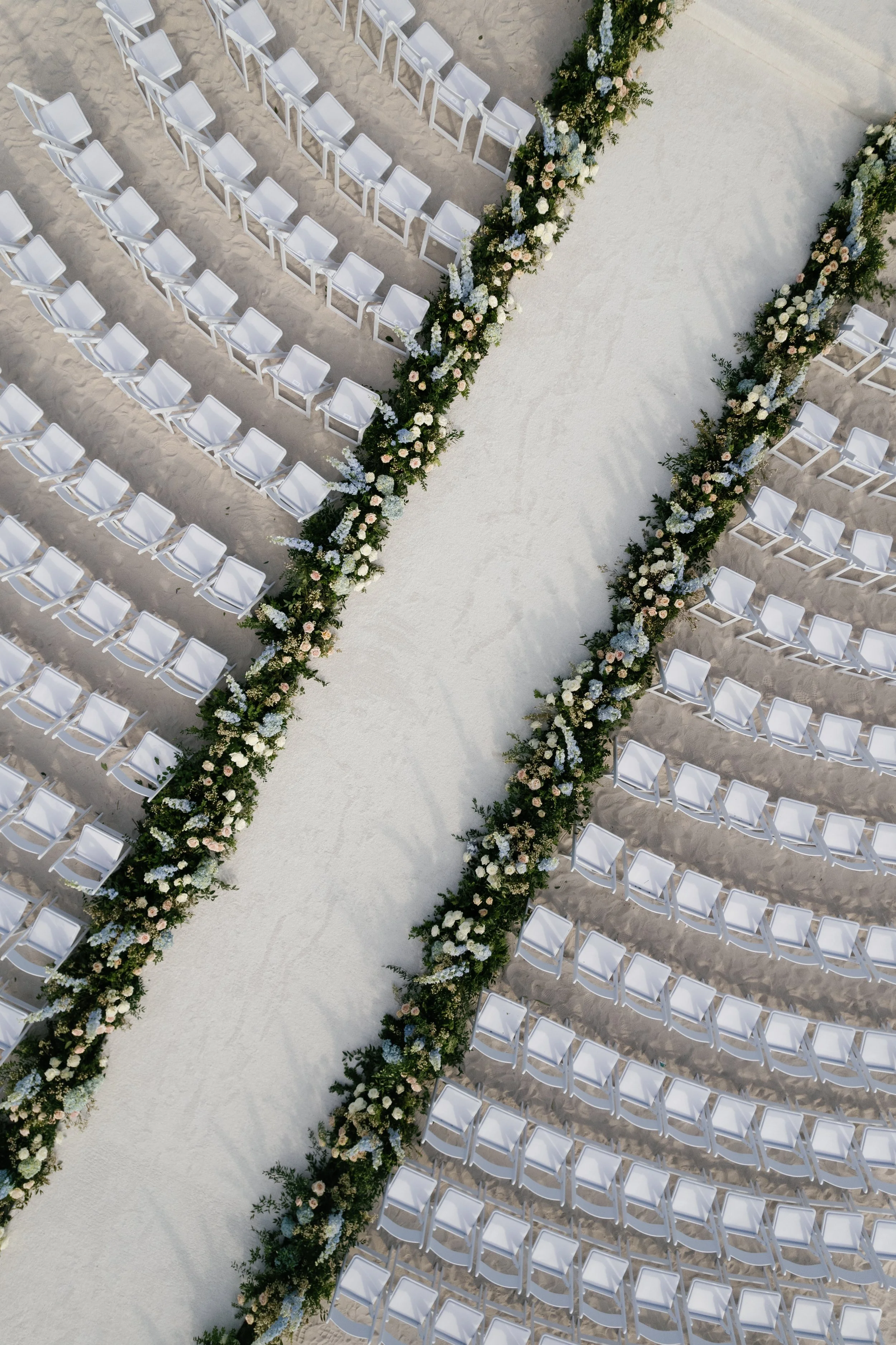 Empty white chairs arranged in rows on sand, separated by a wide aisle decorated with flowers along the edges, set outdoors for a wedding ceremony.