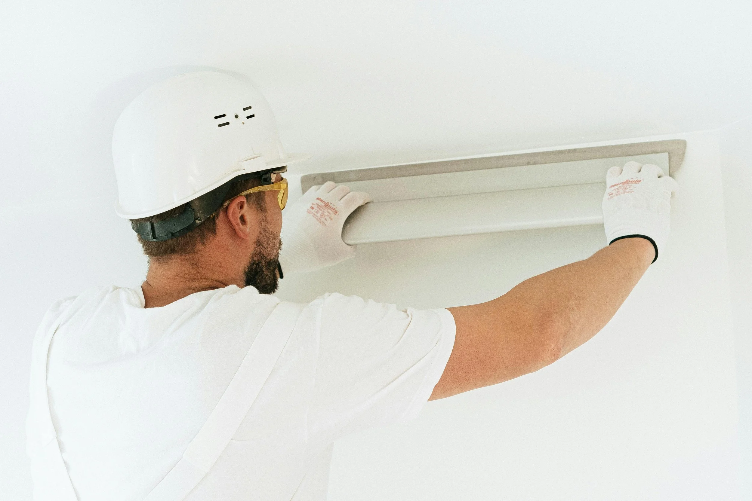 A worker wearing a white helmet, safety glasses, and gloves installing an air vent on a white ceiling.