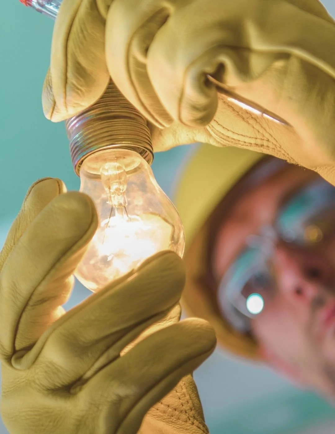 Close-up of a person wearing protective goggles and yellow gloves, installing or inspecting a light bulb with a glowing filament.