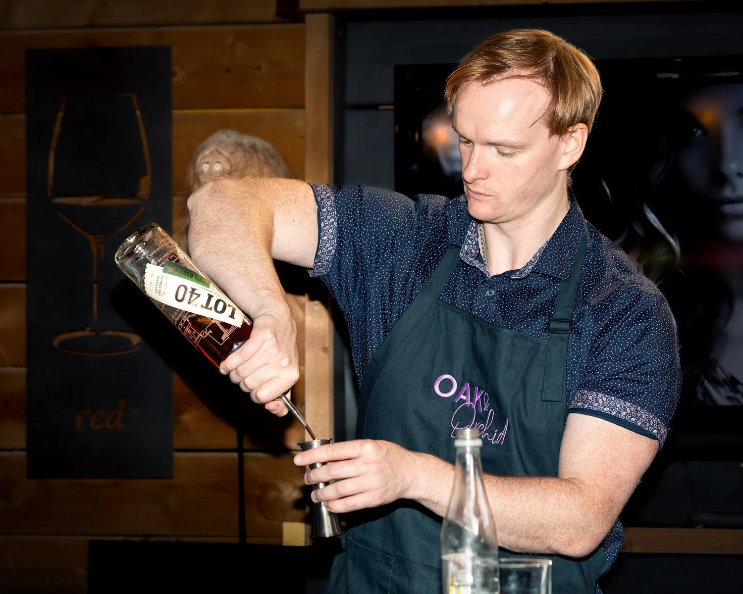 A man pours a dark liquid from a bottle labeled 'Lott 40' into a shot glass. He is wearing a dark apron with purple embroidery that says 'OAK Orchid' and a navy blue, dotted shirt. The background features a wooden wall, and there's a person with blonde hair and glasses behind him.