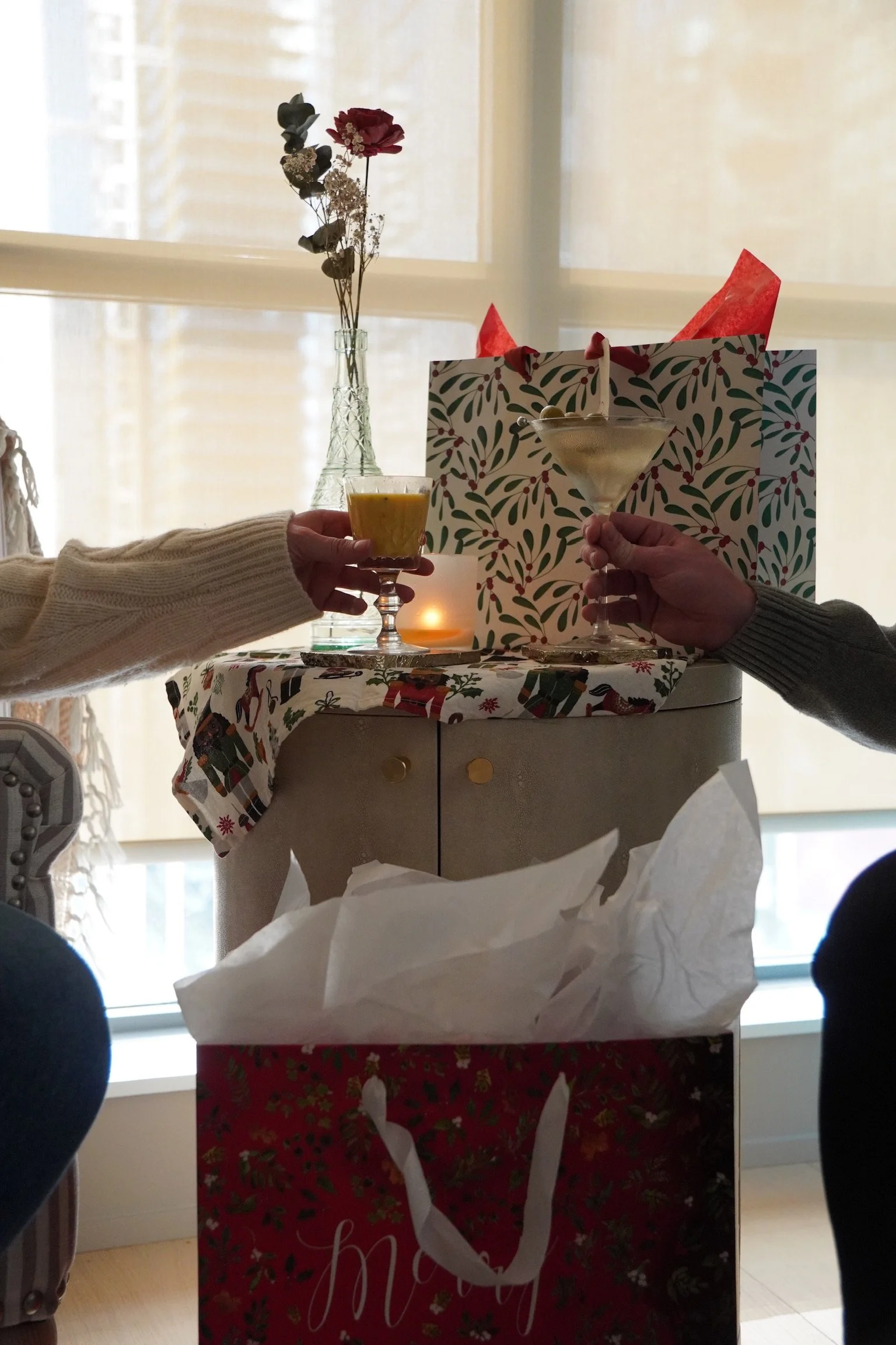 Two people holding drinks, one with a cocktail and the other with a dessert, in front of a holiday-themed gift bag and wrapping paper, with a vase of dried flowers and a lit candle in the background.
