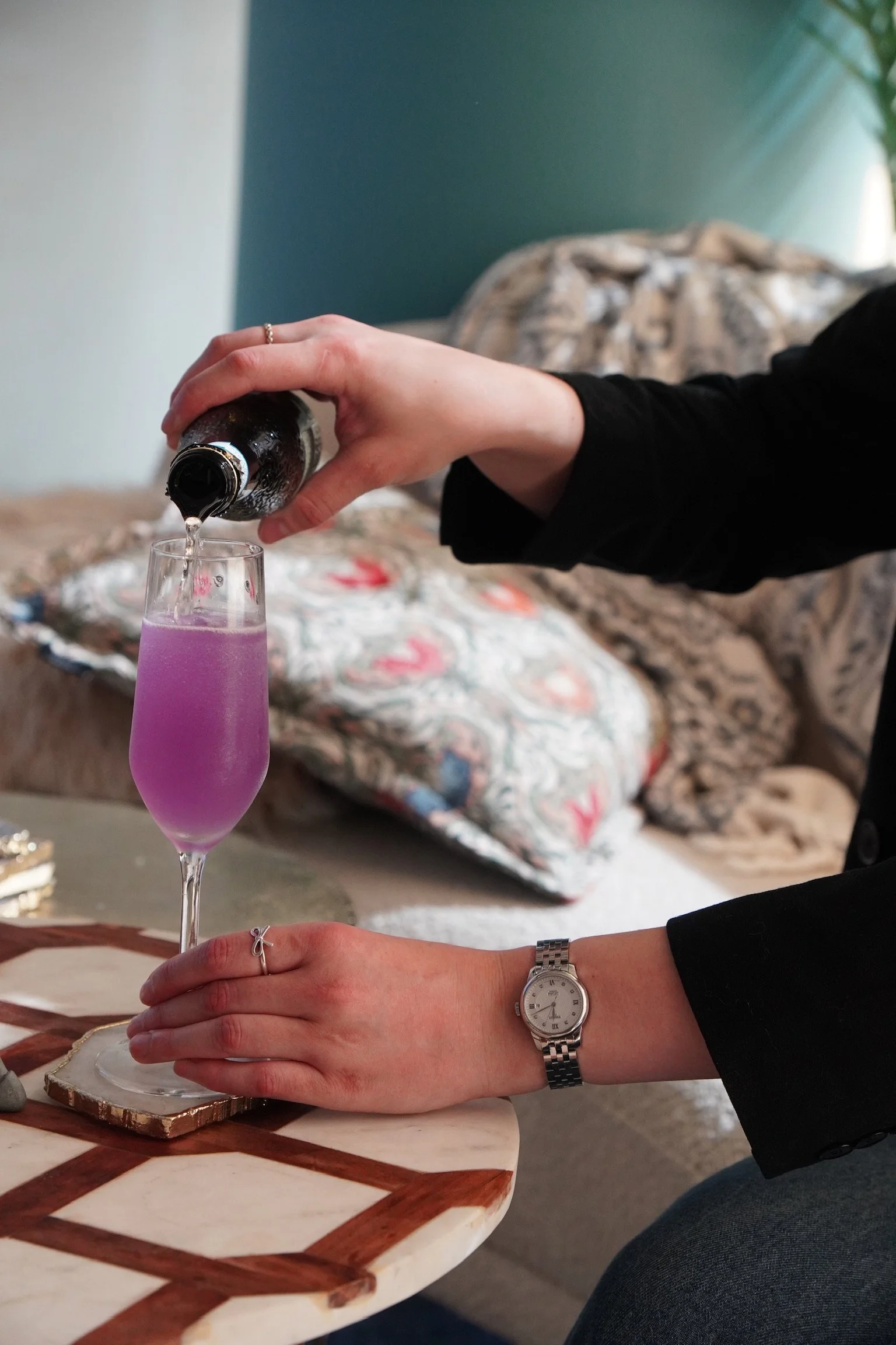 A person is pouring a purple drink into a champagne flute from a dark bottle, with their left hand resting on the table. They are wearing a silver watch and a ring, and are seated in a room with patterned pillows and a green wall in the background.