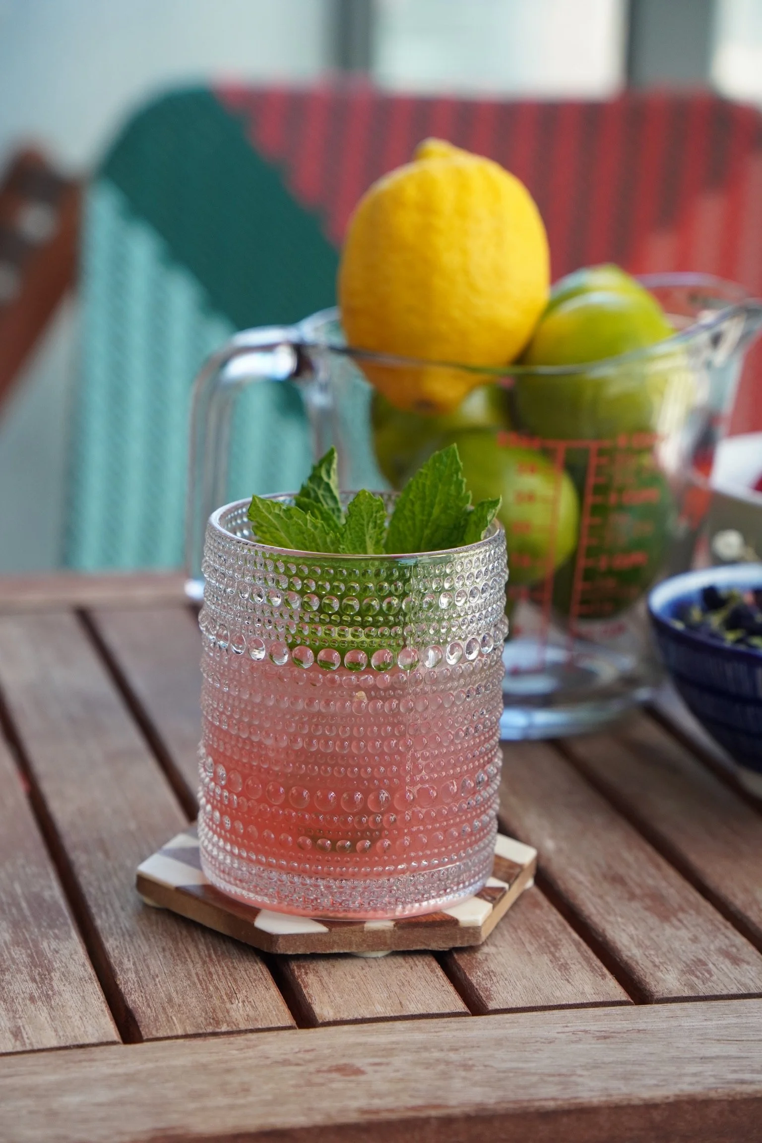 A pink cocktail with mint leaves in a textured glass on a wooden table, with a pitcher of lemons and green apples in the background.