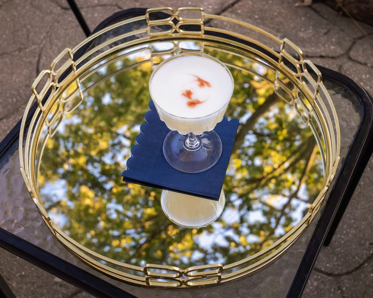 A cocktail glass filled with a white frothy drink garnished with red leaves, placed on a blue napkin on a mirrored surface. The mirror reflects a tree with yellow and green leaves.
