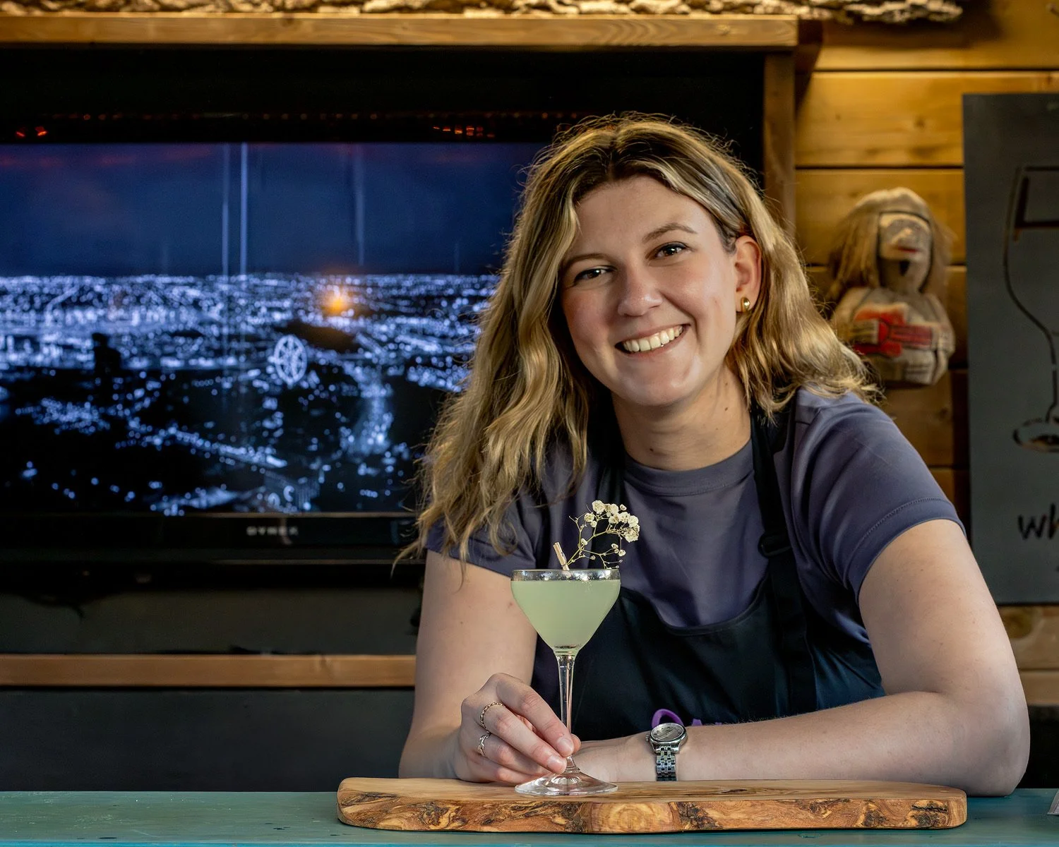 A woman with blonde hair smiling while sitting at a bar, holding a green cocktail with a garnish, in a setting with wooden walls and a large screen displaying a night cityscape in the background.