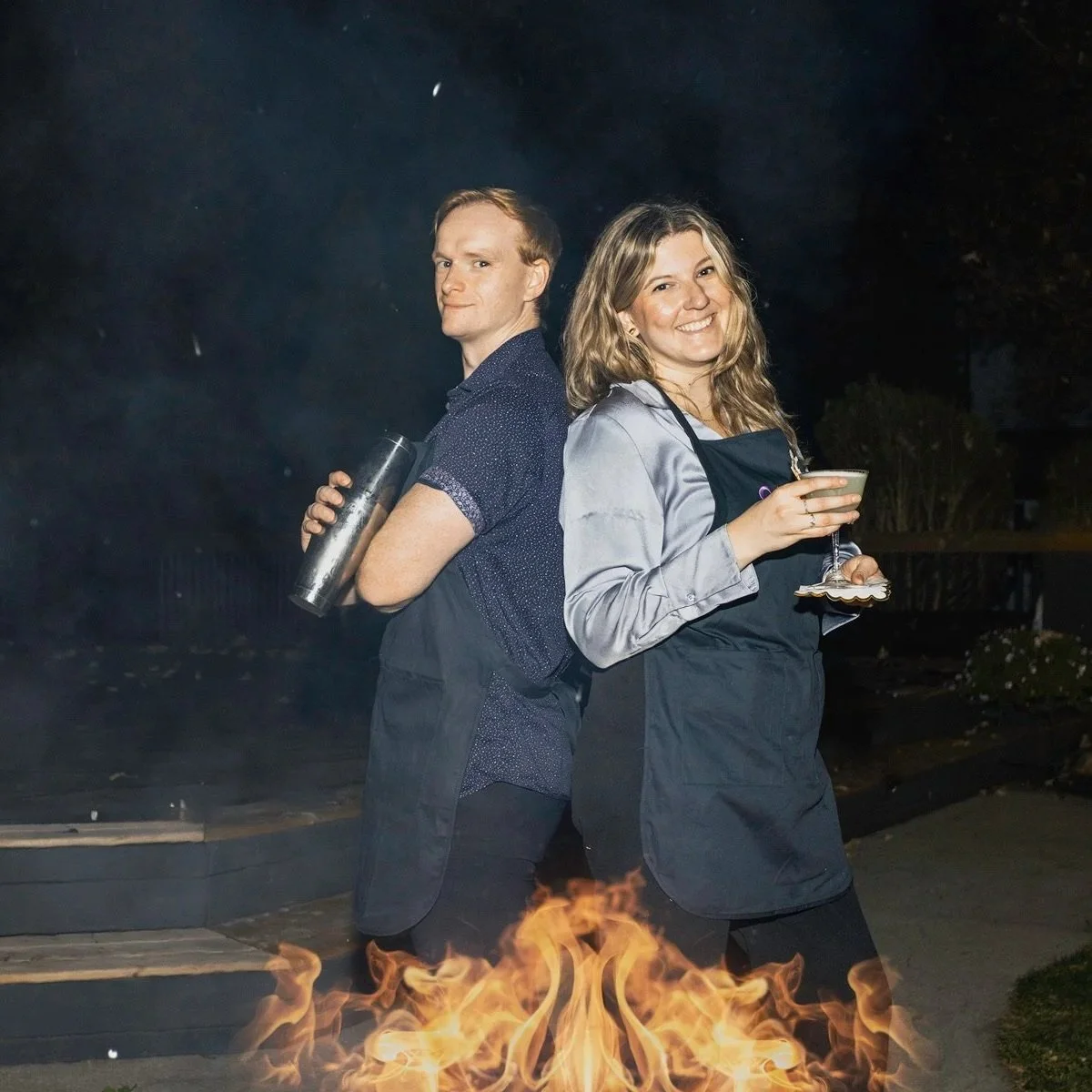 Two people, a man and a woman, standing back to back outdoors at night with a fire in front of them. The man is holding a tumbler and the woman has a cocktail glass. Both are wearing aprons and smiling or making a face.