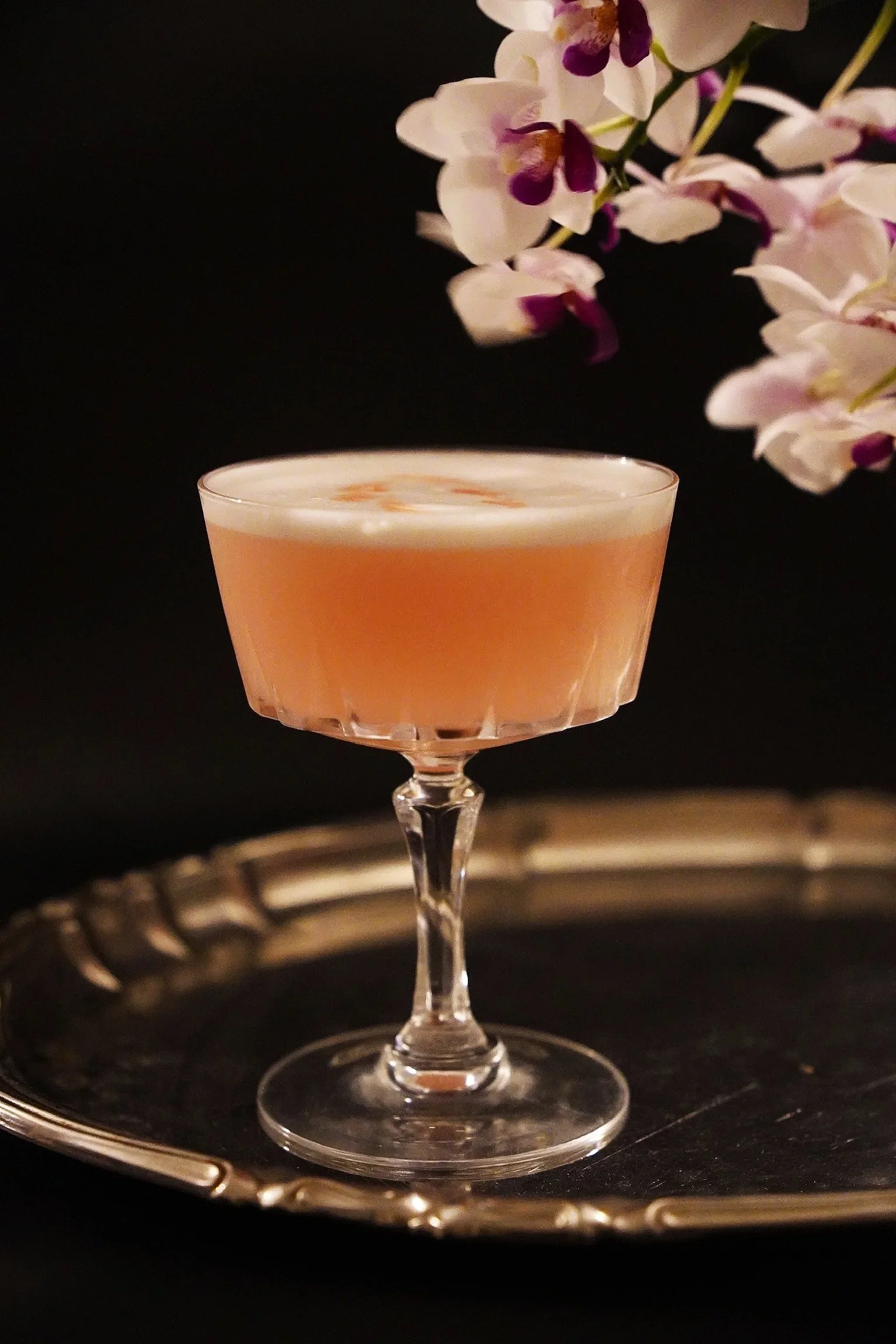A pink cocktail in a vintage coupe glass placed on a silver tray, with purple and white orchids in the background against a dark backdrop.