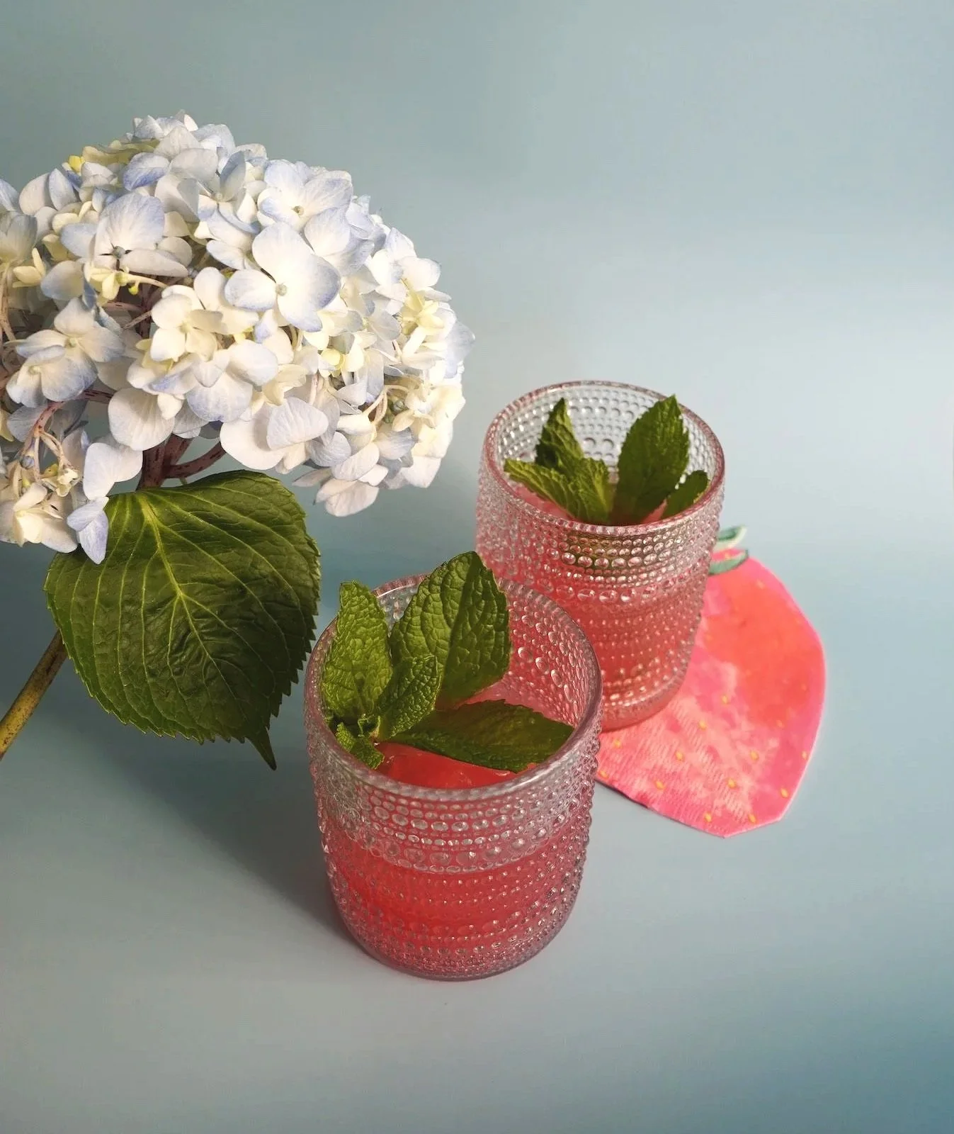 Two pink glasses with textured surfaces filled with pink drink and mint leaves, placed on a pink napkin, with white hydrangea flowers and green leaves on a light blue background.