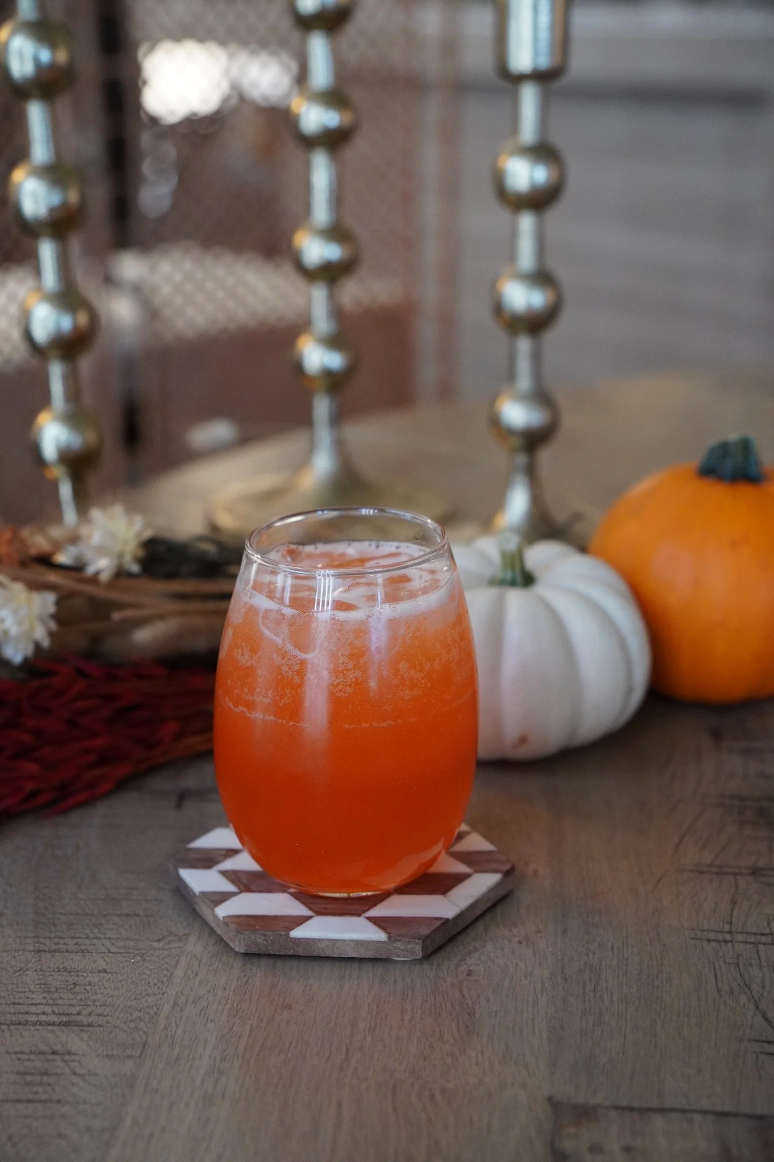 A glass of orange-colored beverage on a wooden table, with white and orange pumpkins and decorative balls in the background.