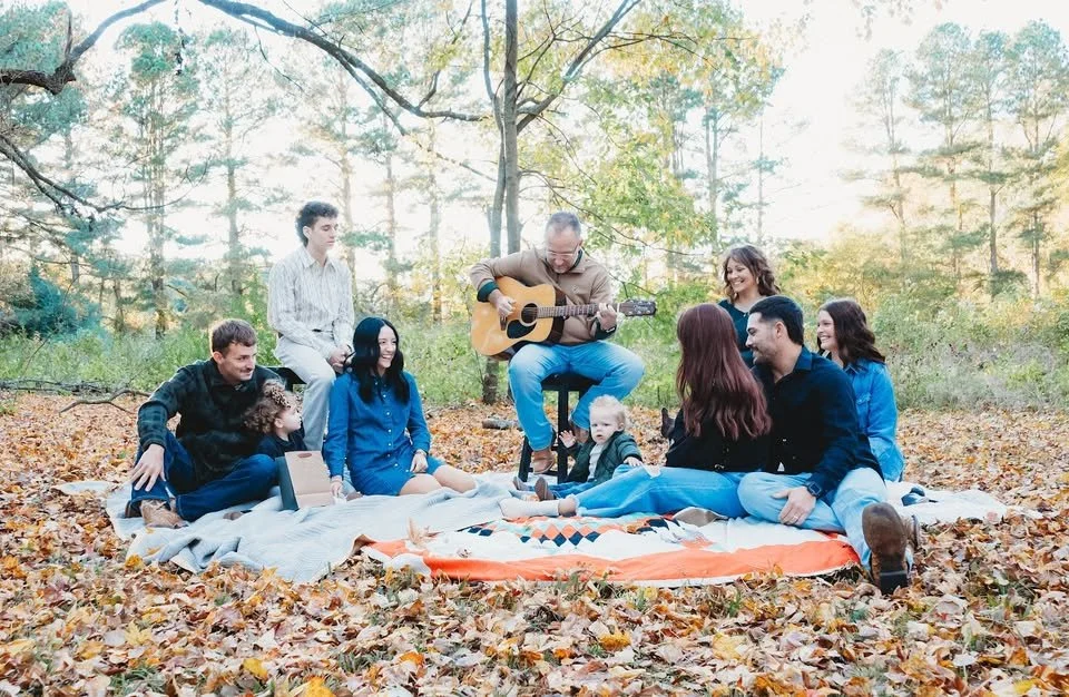Family and friends enjoying an outdoor picnic in a forest, with a man playing guitar surrounded by children and adults sitting on blankets.