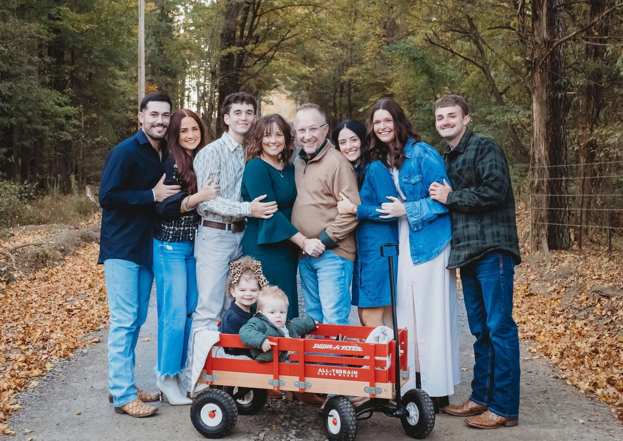 A group of eight adults and two children standing outdoors on a leaf-covered trail in a wooded area during fall, smiling and posing for a photo.