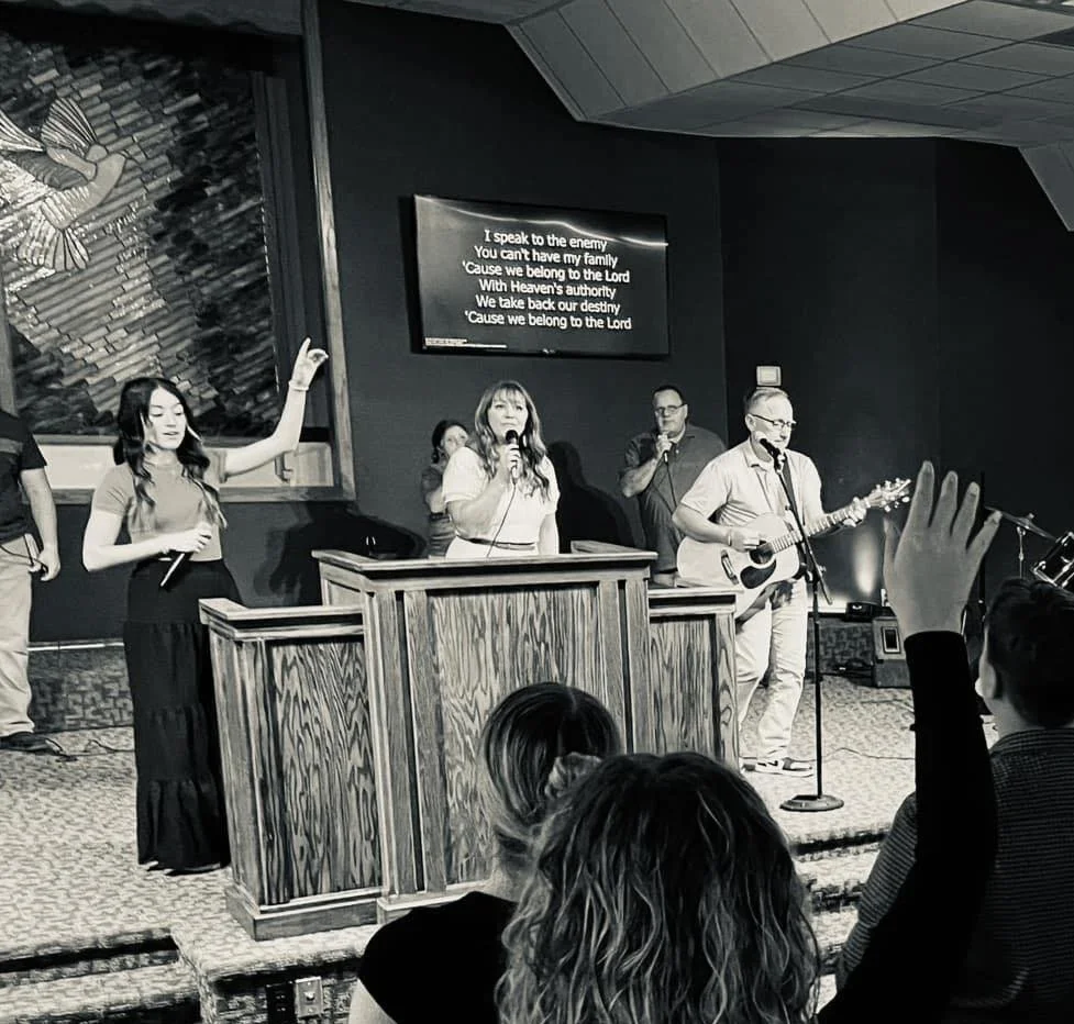 A church service with a band performing on stage, including a woman singing and a man playing guitar, with several people in the audience raising their hands.