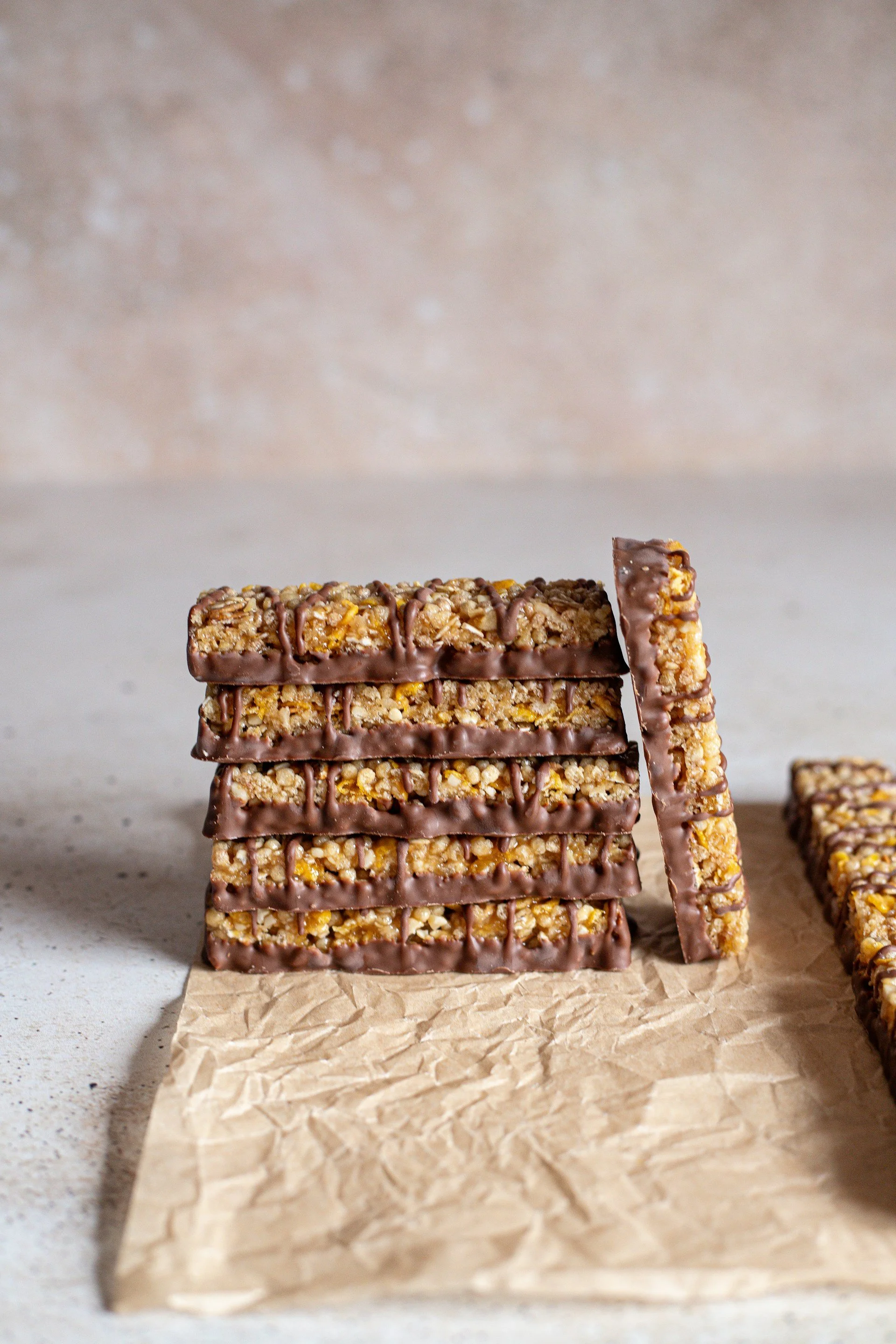 Stacked granola bars with chocolate coating on a piece of crumpled brown parchment paper, with a blurred neutral background.