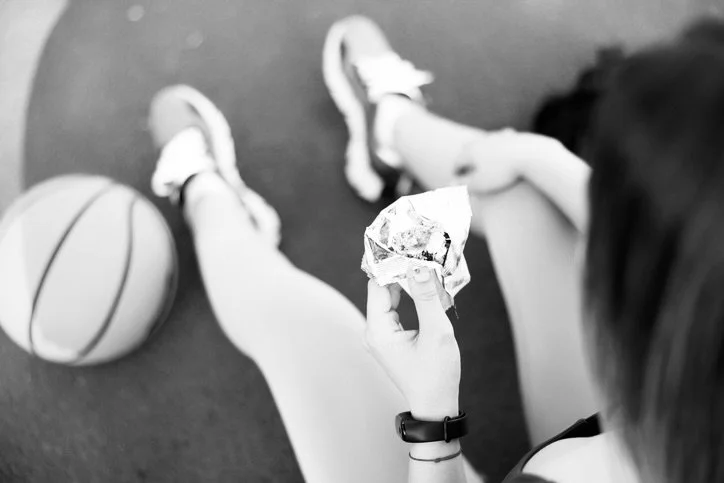 A person sitting on the floor holding a wrapped sandwich, with a basketball nearby and wearing athletic shoes and a wristwatch.