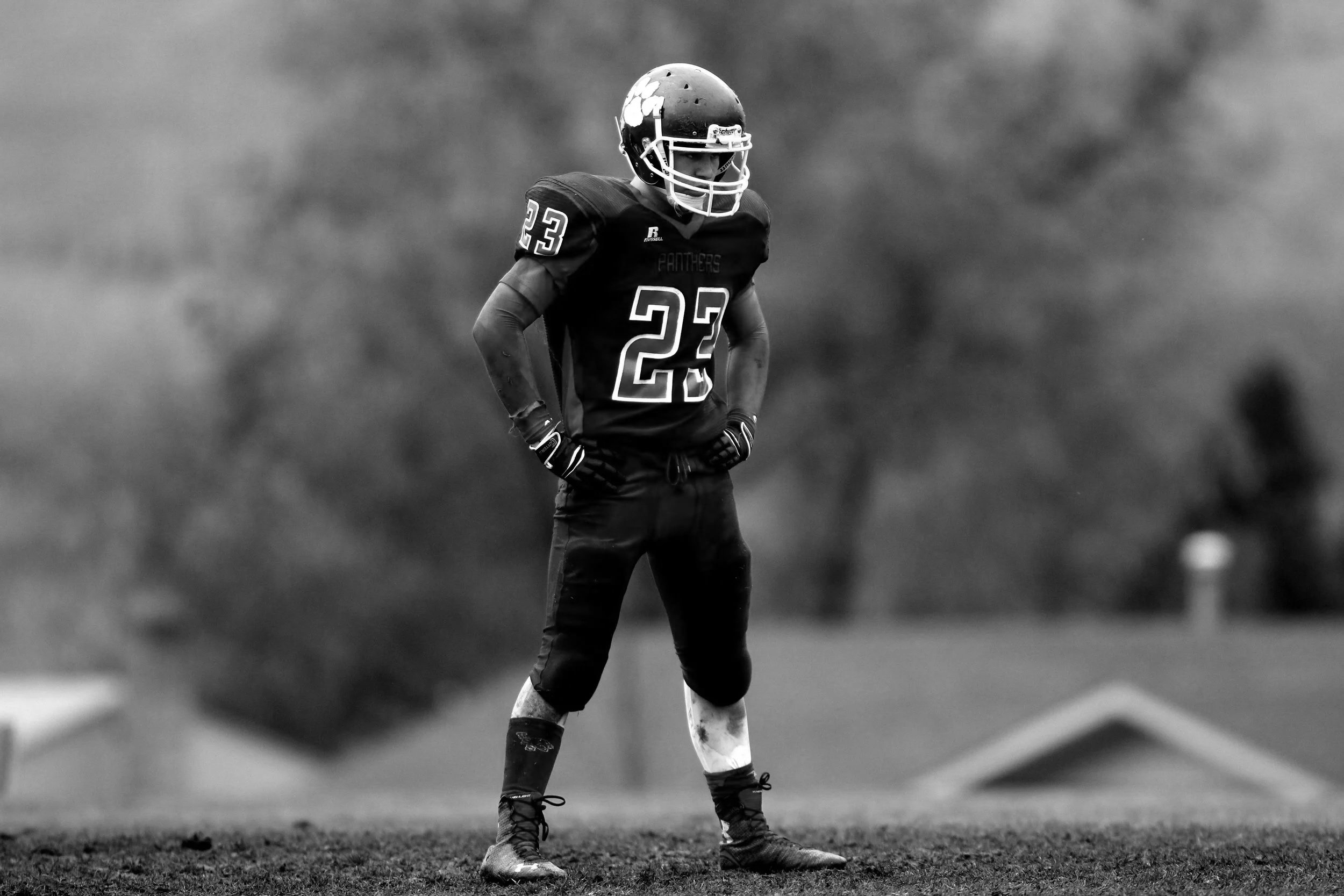 A young football player stands on the field in a black uniform with the number 23, wearing a helmet with a face mask, gloves, and cleats, outdoors on a cloudy day.