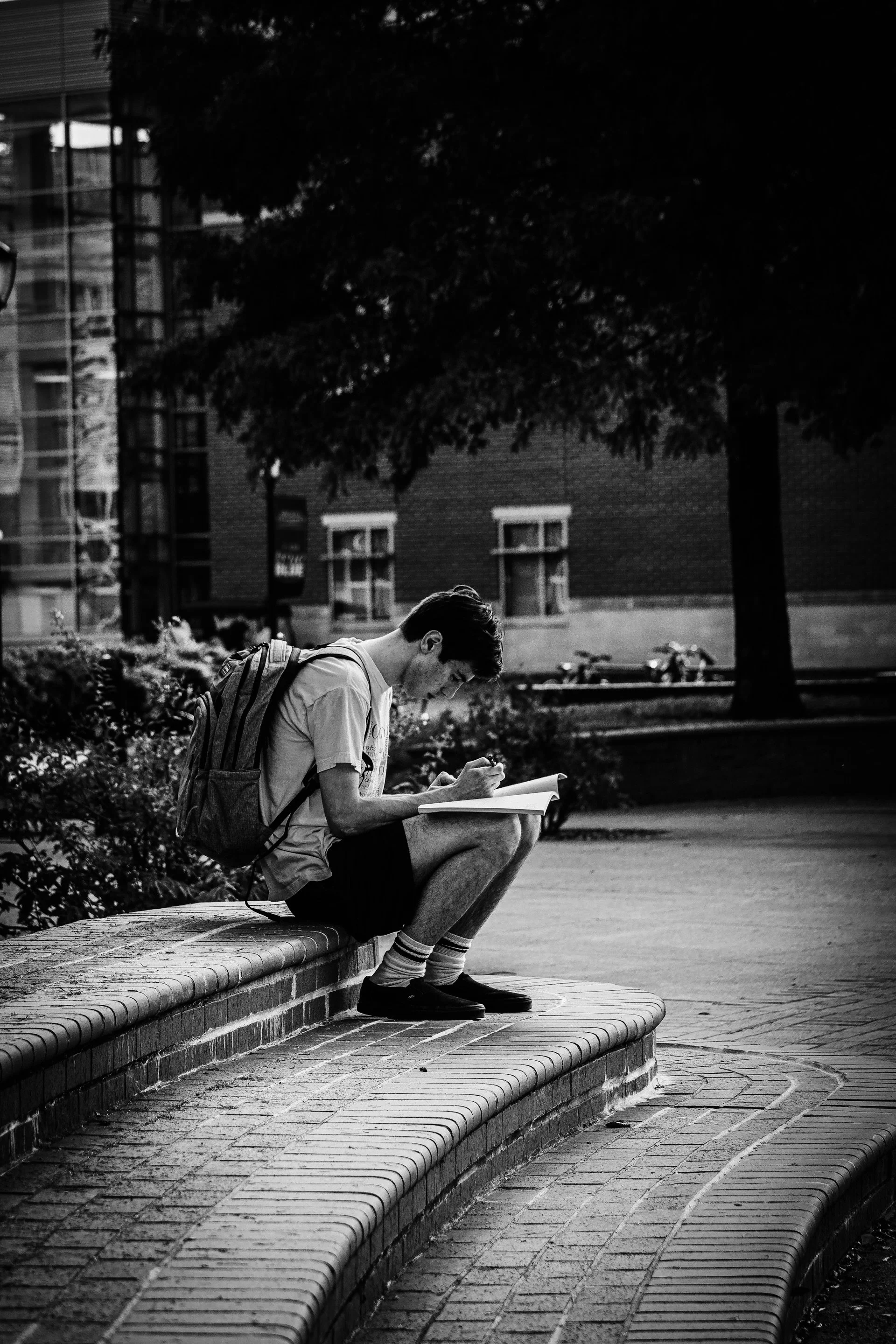 A young man sitting on a curved brick bench outdoors, writing in a notebook with a backpack on his back, surrounded by trees and buildings.