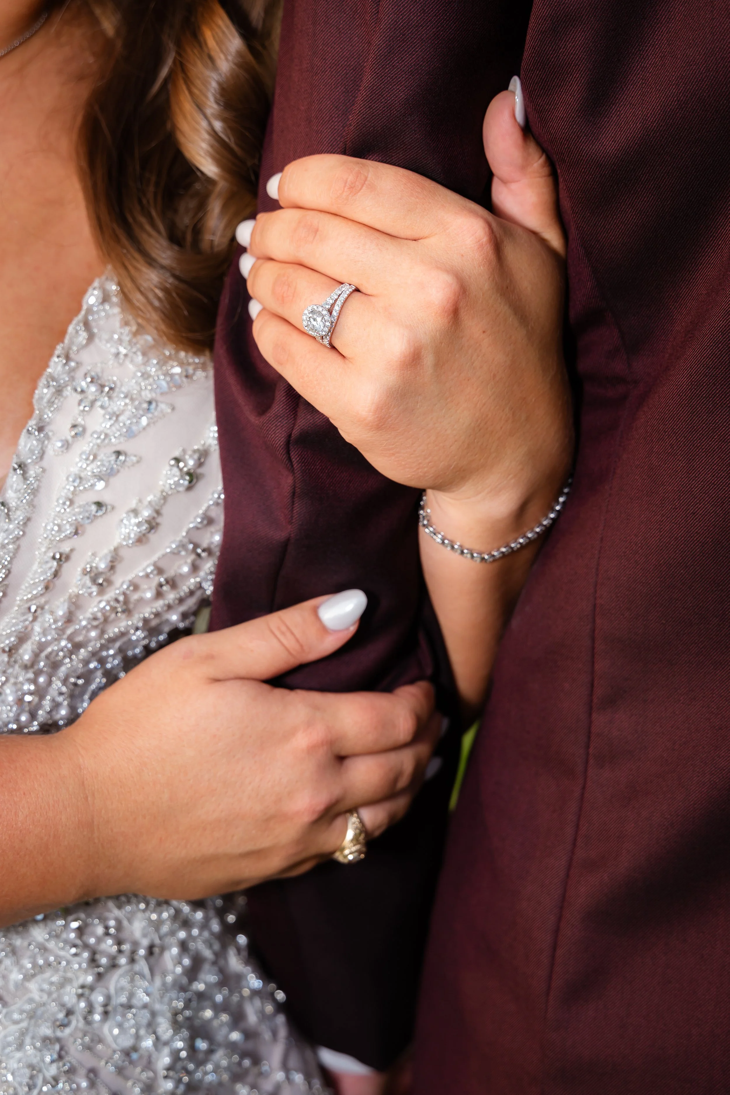 Close-up of a couple holding each other's arms, with the woman's wedding ring visible on her left hand, which is placed on the man's arm. The woman is wearing an ornate, beaded dress and a bracelet, and is holding onto the man's arm with her other ha