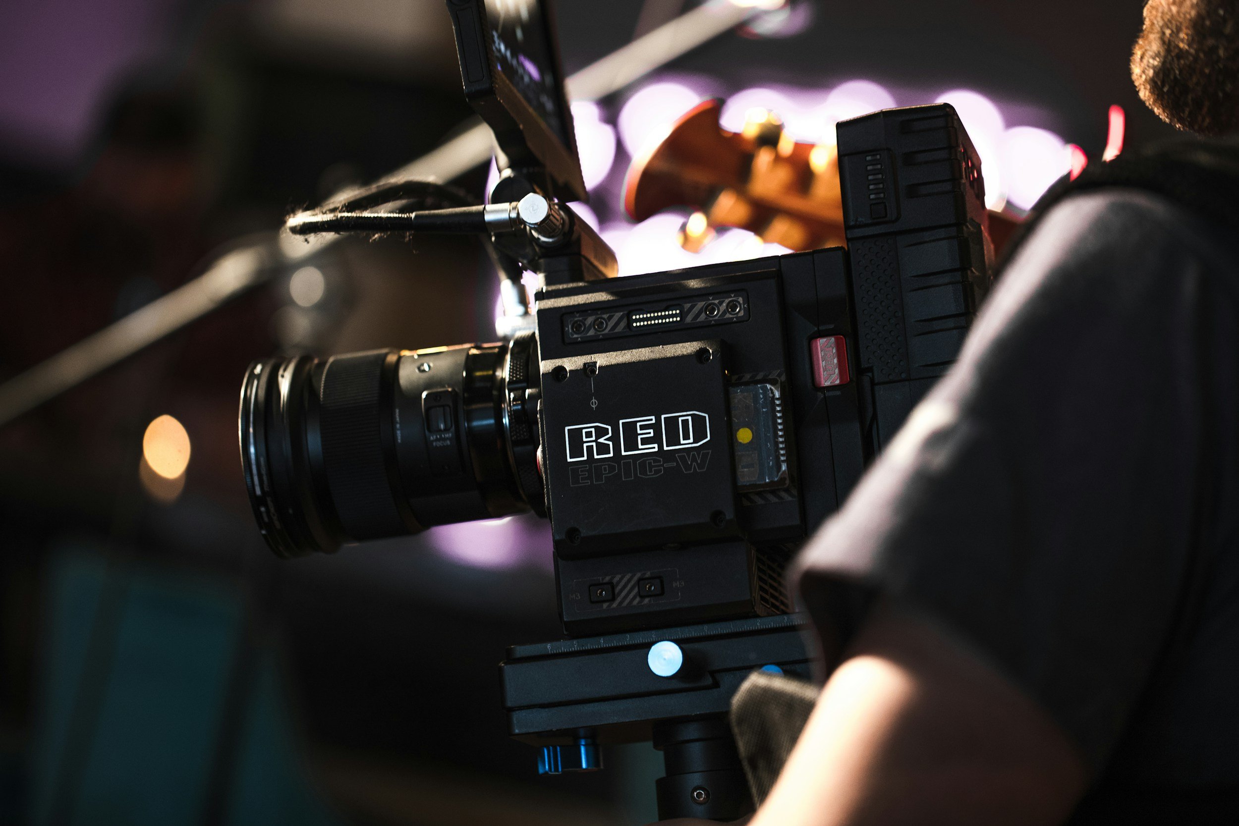 Close-up of a professional film or TV camera setup with a large lens, marked 'RED EPIC-W,' on a tripod in a production environment with blurred bright lights in the background.