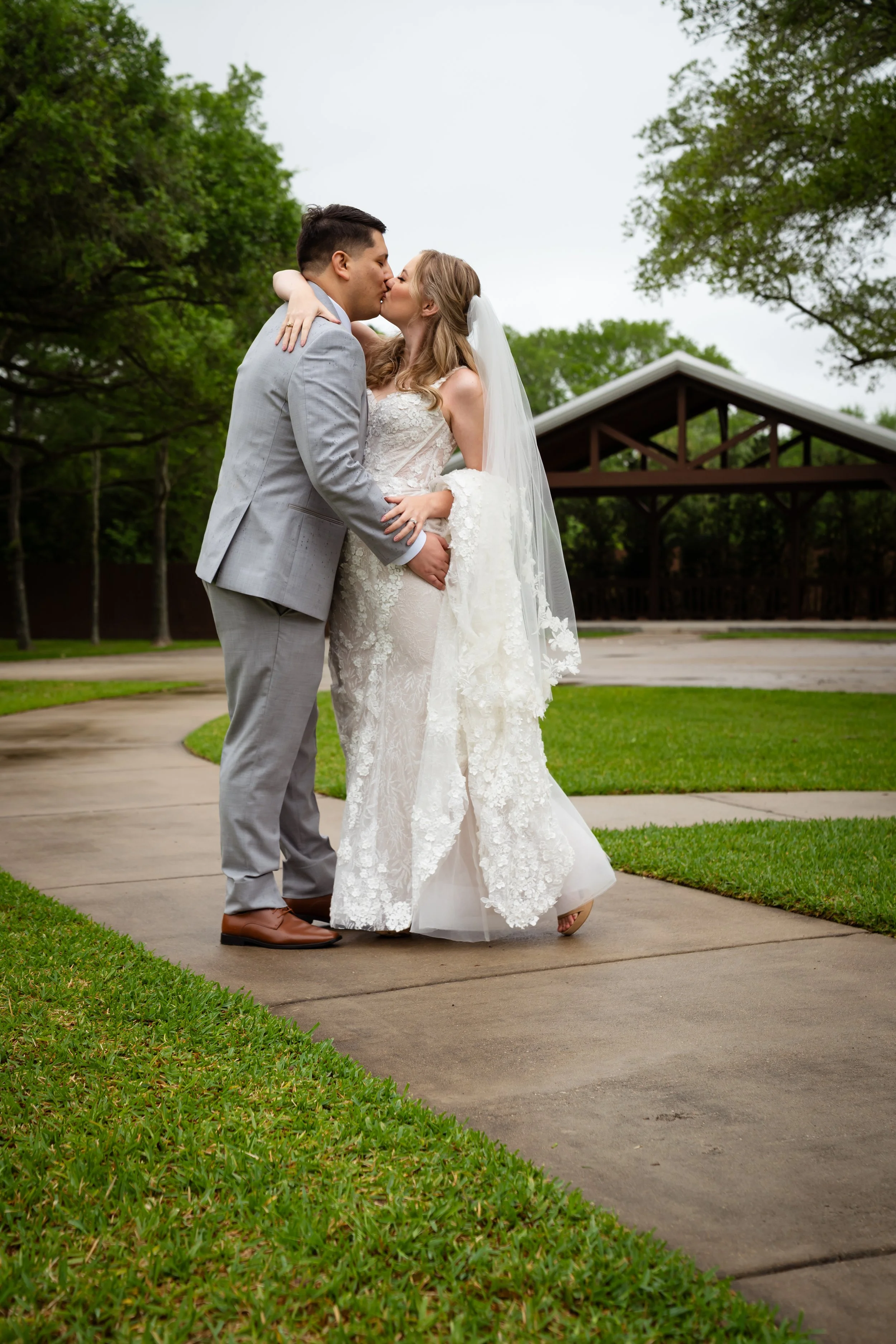 A bride and groom kissing outdoors on a cloudy day, with green trees and a pavilion in the background.