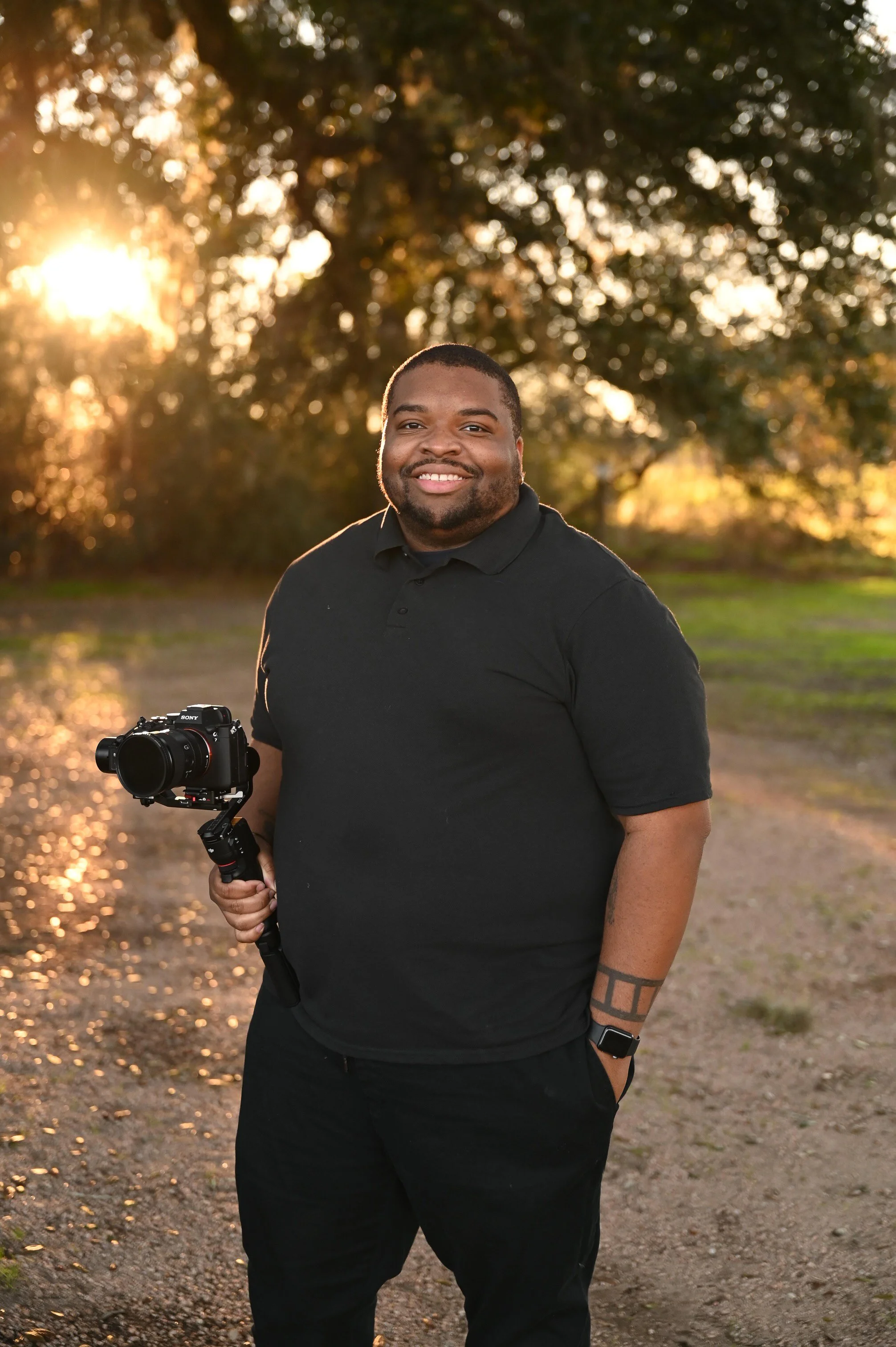 A smiling man holding a camera outdoors during sunset, standing on a dirt path with trees in the background.