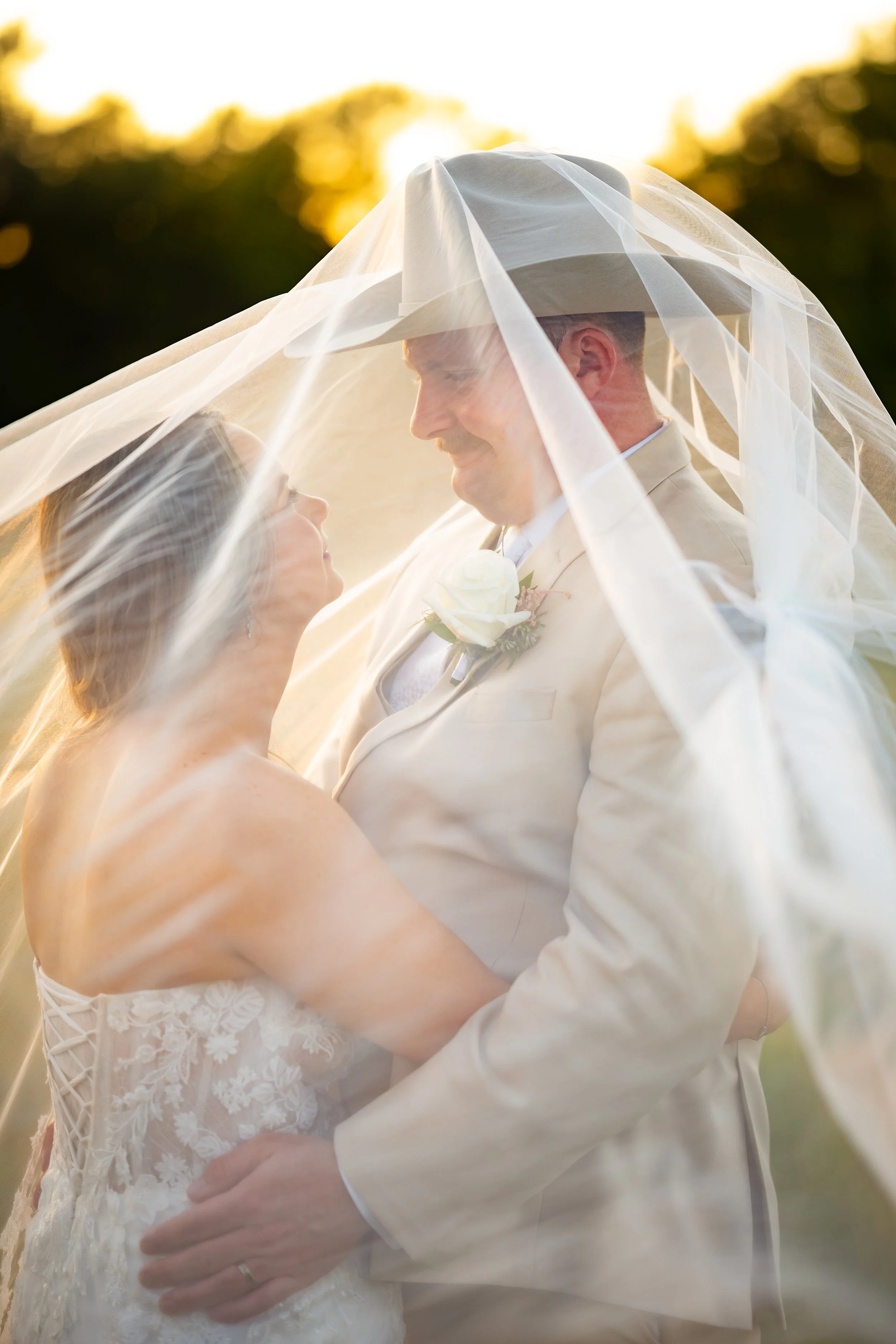 A bride and groom on their wedding day, smiling and gazing at each other under a veil, with the sun setting behind them.