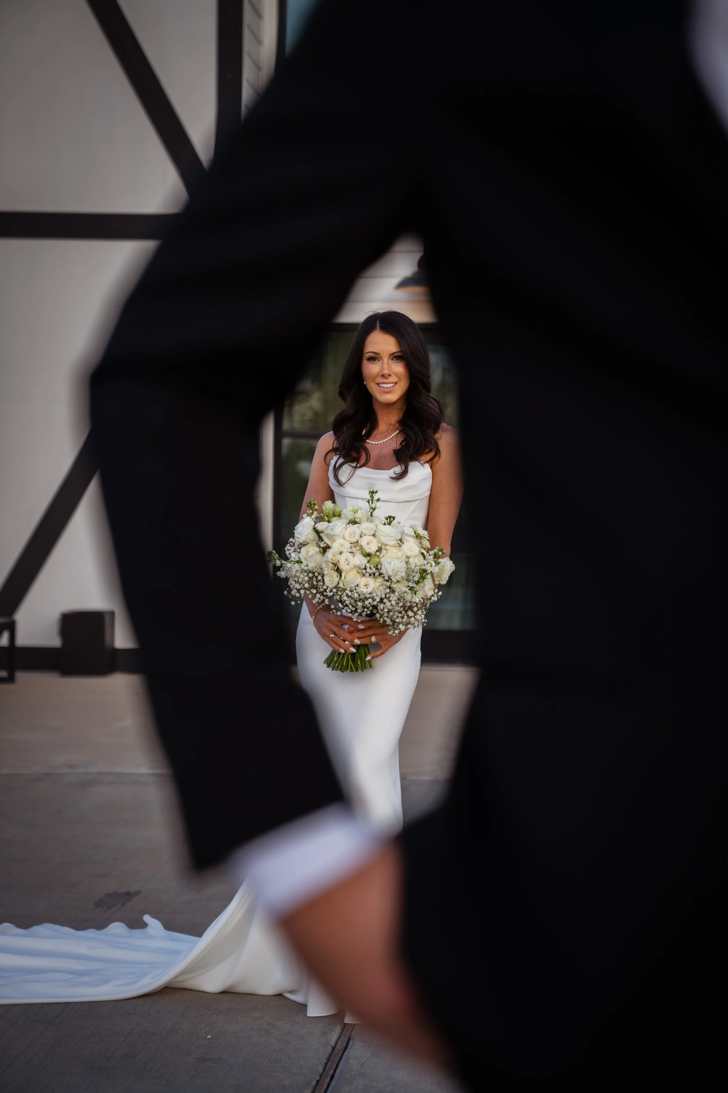 A bride with dark hair in a white wedding dress holding a bouquet of white roses, framed by a person's arm and a window in the background.