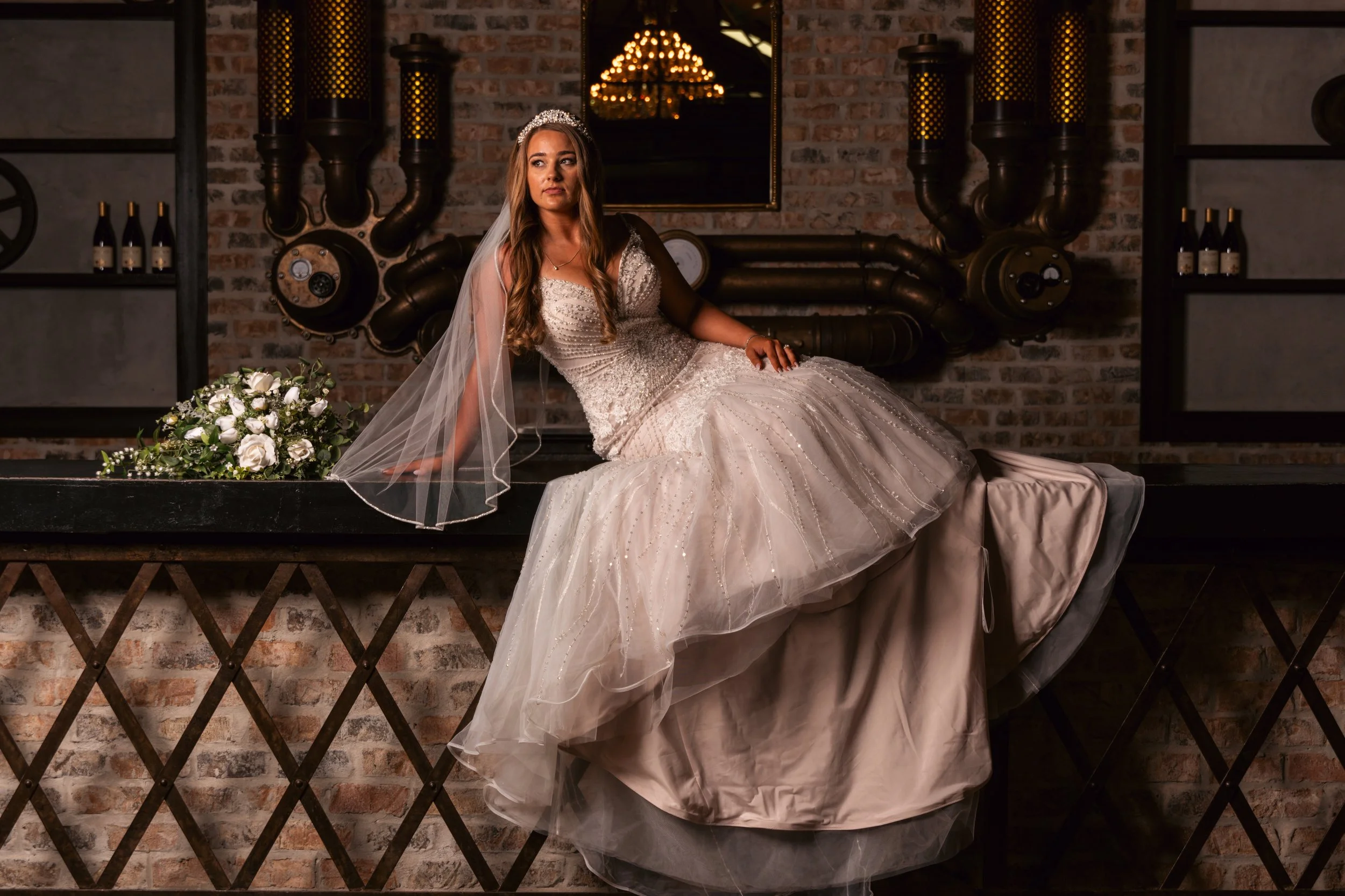 A woman in a wedding dress sitting on a black ledge in front of a brick wall and industrial pipes, with a bouquet of white flowers beside her.
