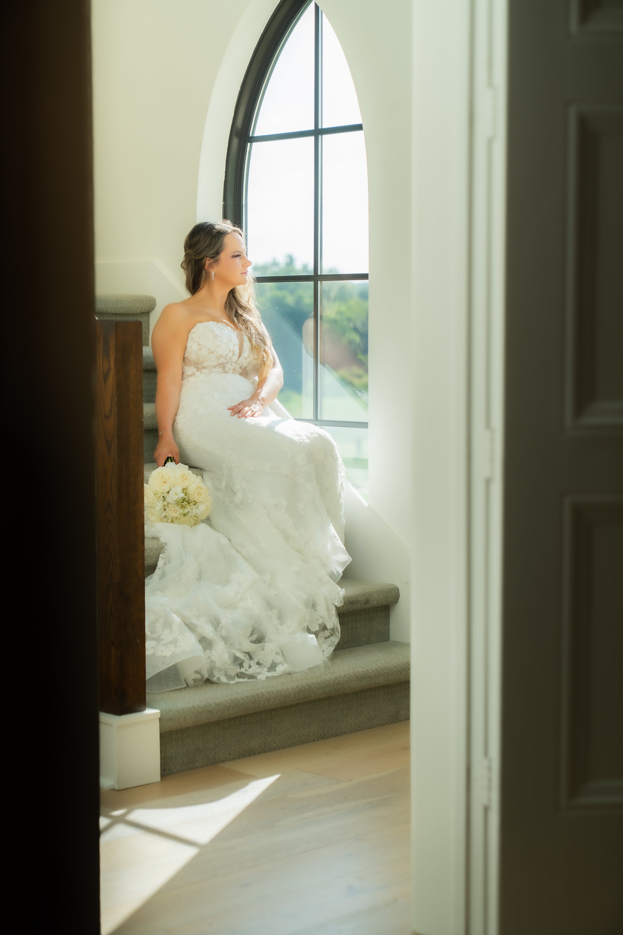A bride sitting on stairs beside a large arched window, holding a bouquet of white roses, illuminated by natural light, wearing a strapless lace wedding gown.