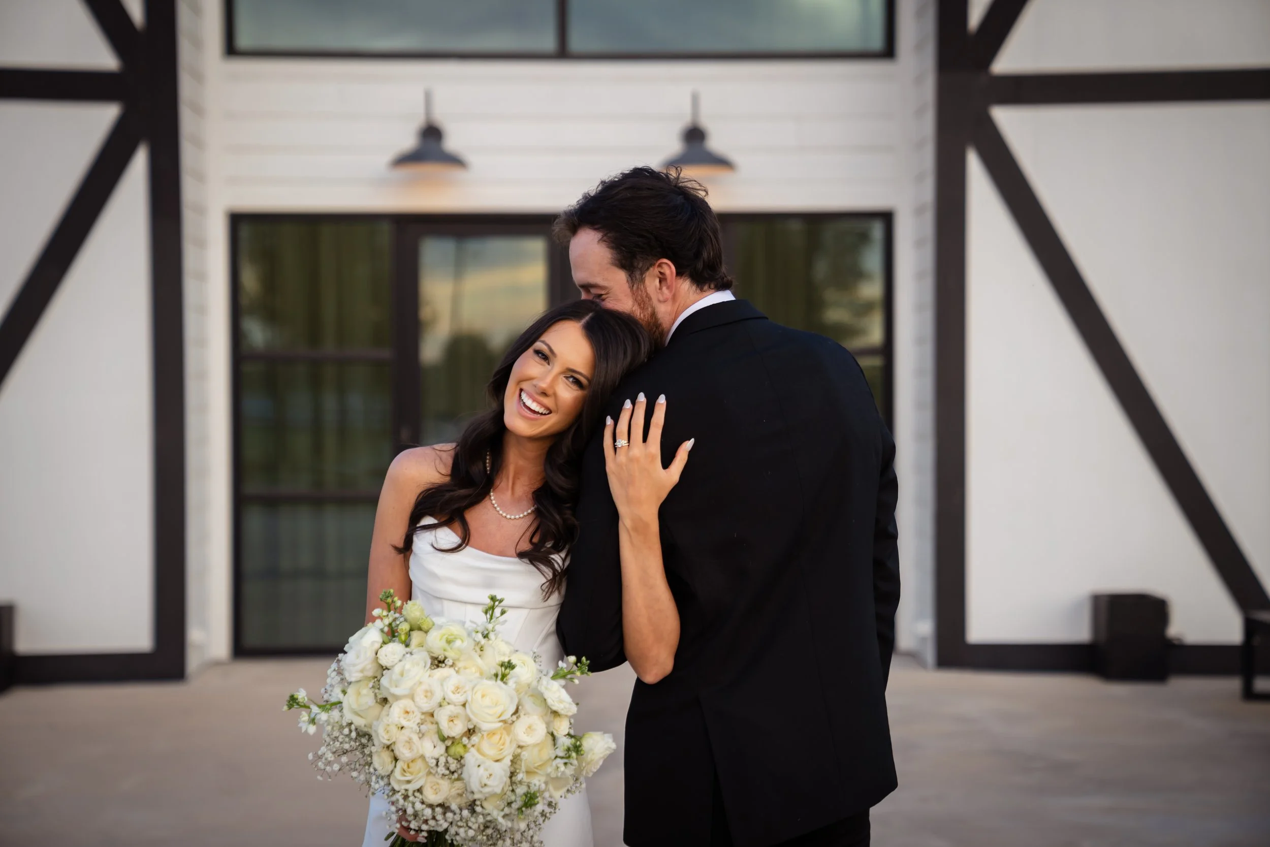 A bride and groom smiling and embracing on their wedding day outside a modern building with black trim and three hanging lights.