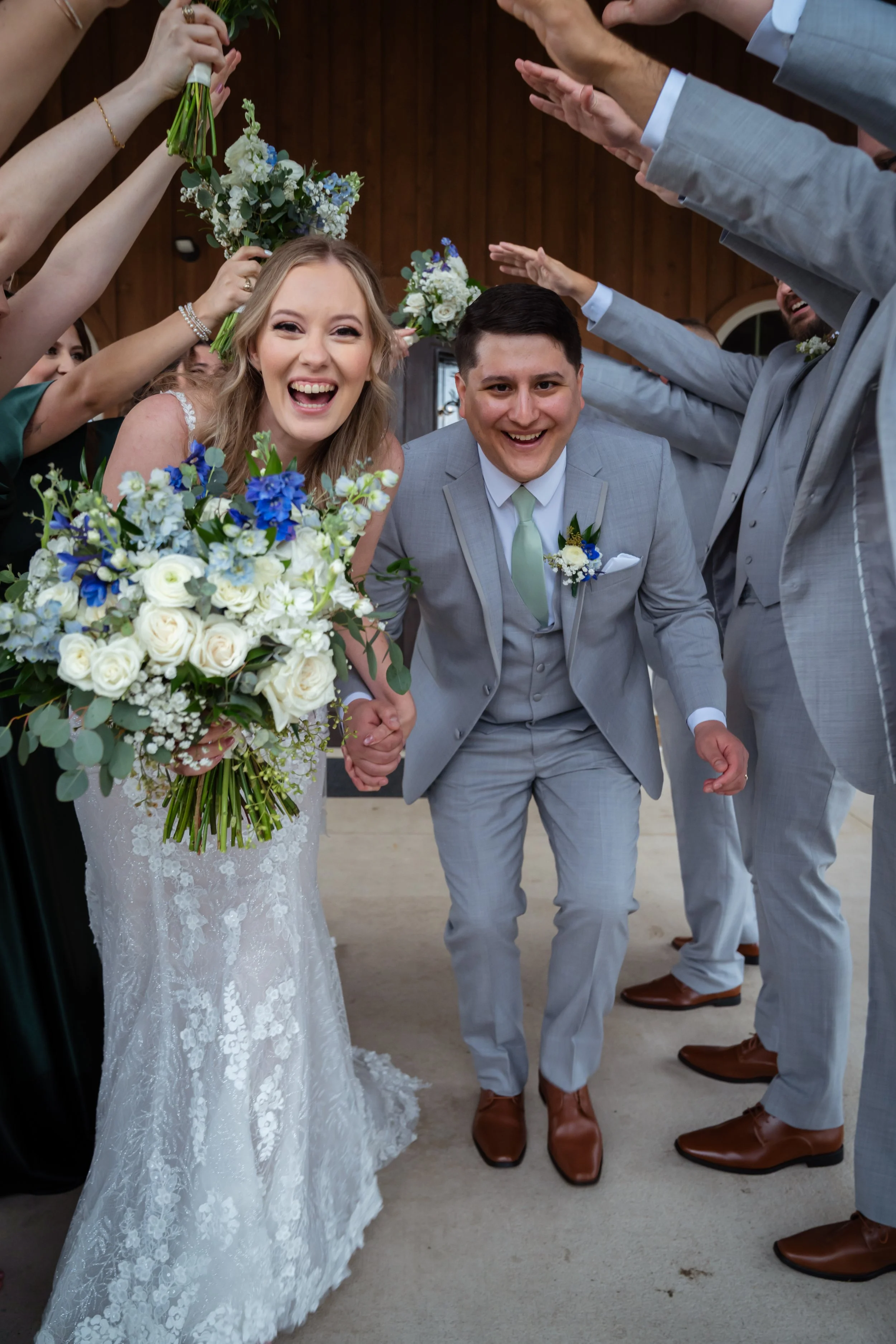A newlywed couple holding hands and smiling, surrounded by friends and family at a wedding celebration. The bride holds a large bouquet of white roses and blue flowers, and wears a lace wedding dress. The groom wears a light gray suit with a mint gre