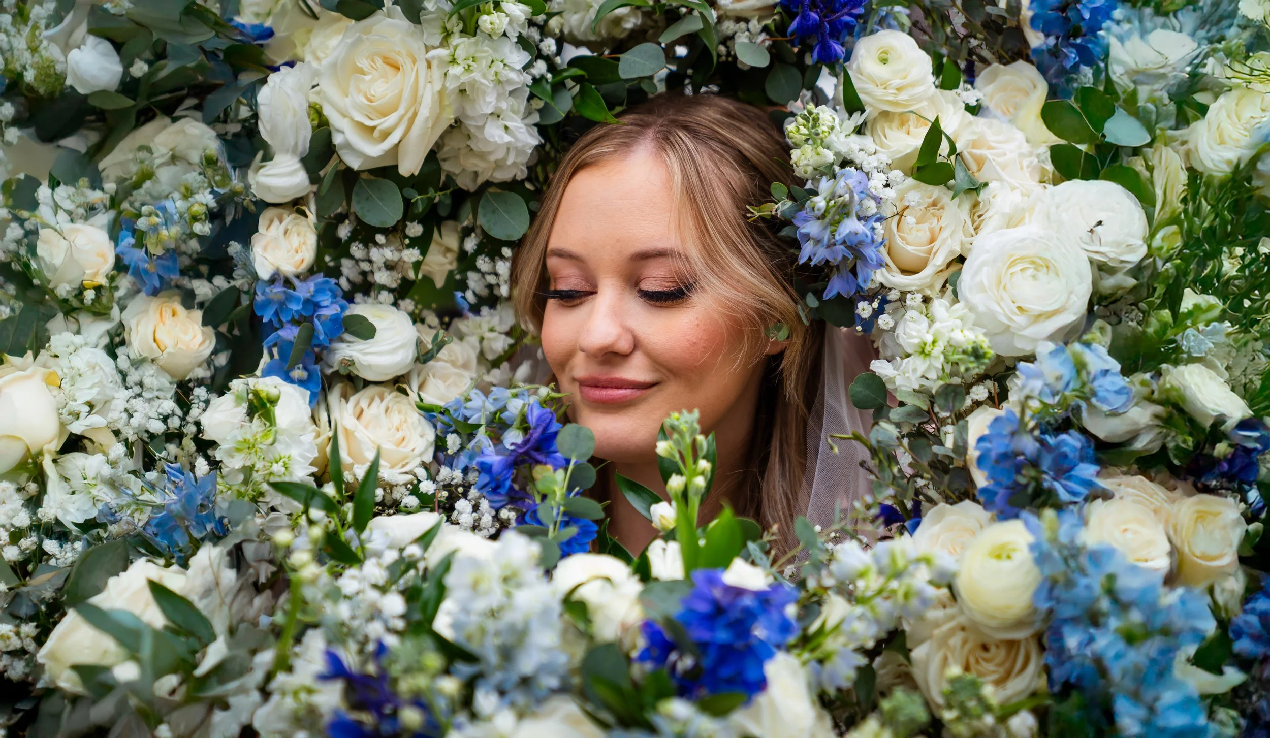 A bride with closed eyes surrounded by a lush arrangement of white, cream, and blue flowers.
