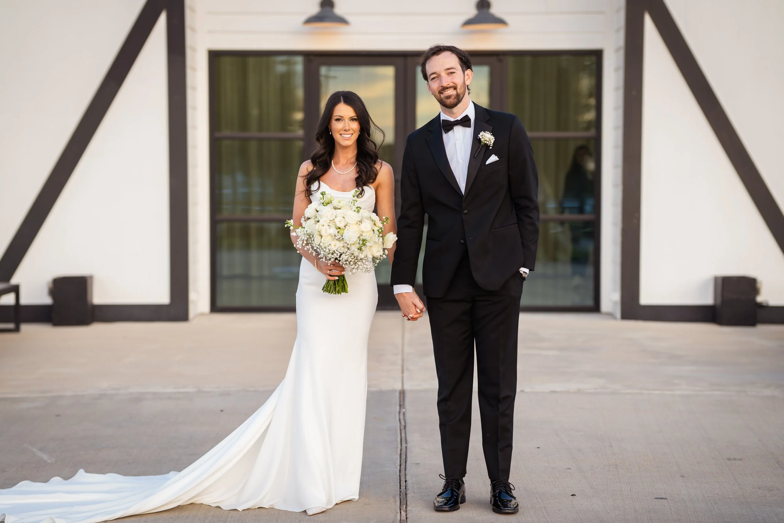 A newlywed couple standing outdoors, holding hands, with the bride holding a bouquet of white flowers and wearing a white wedding dress, and the groom in a black tuxedo with a bow tie, smiling.