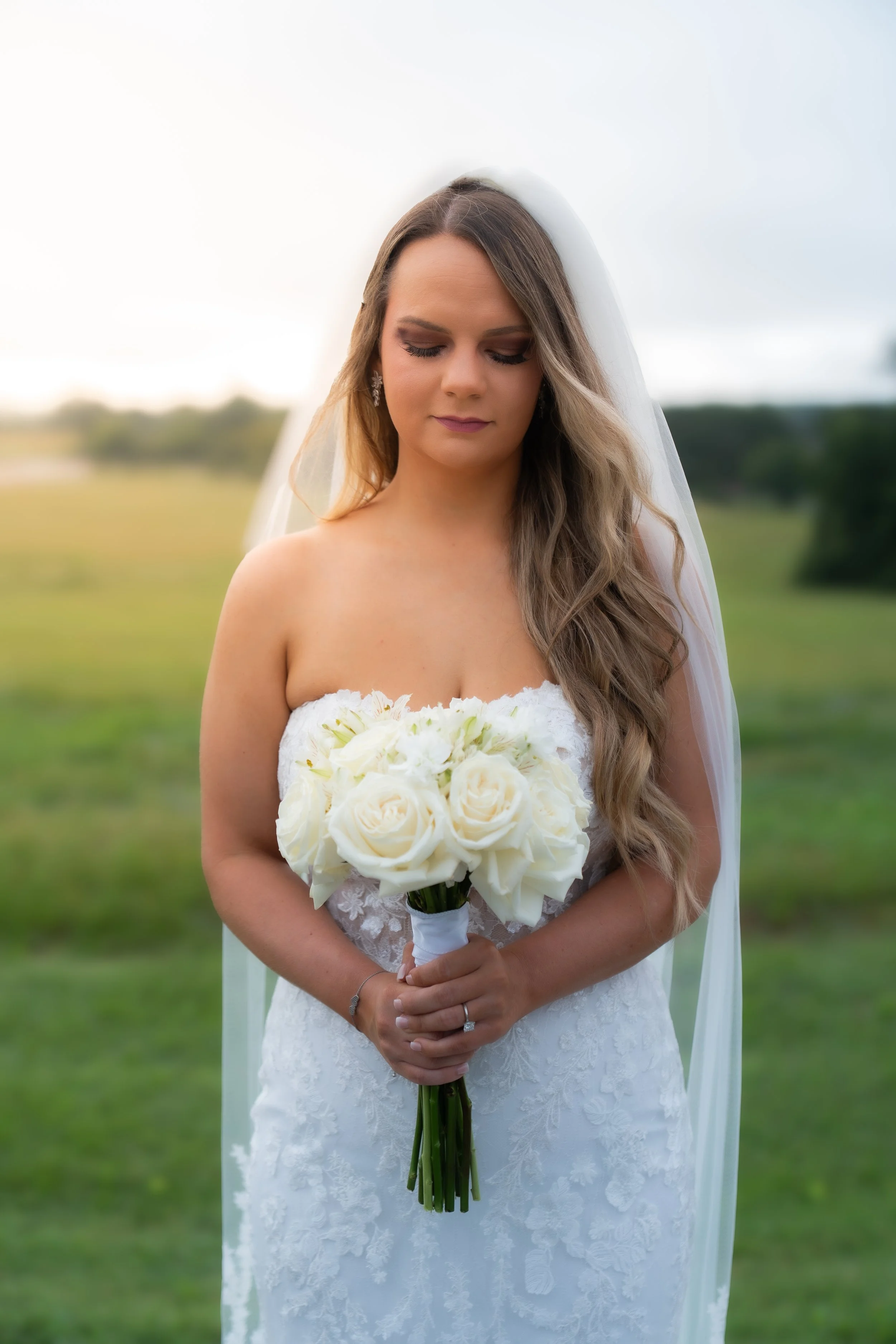 A bride in a strapless white wedding dress holding a bouquet of white roses standing outdoors with a blurred green landscape background.
