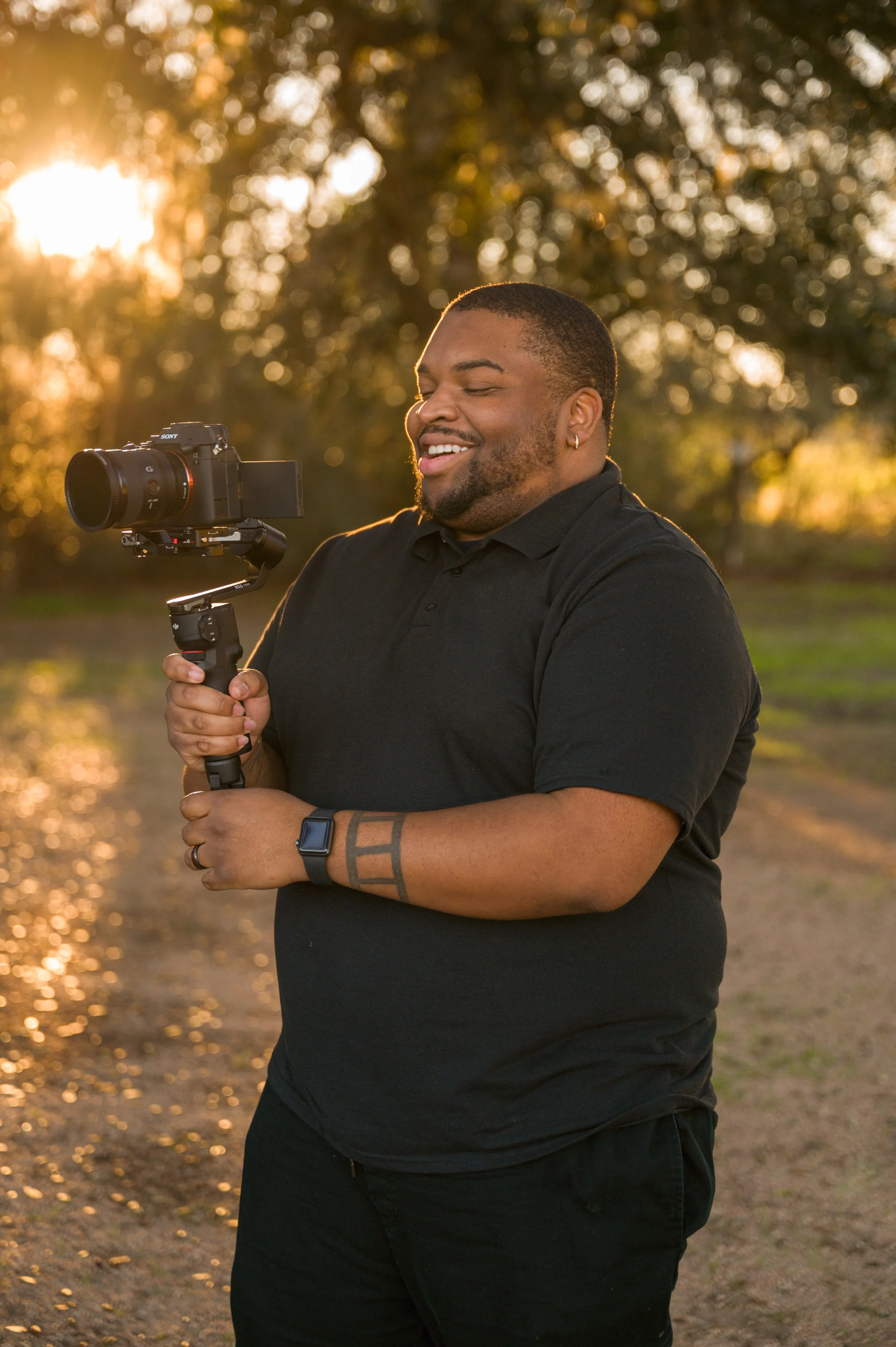A man with dark hair, beard, and earrings, wearing a black polo shirt and smartwatch, is smiling while holding a gimbal-stabilized camera outdoors during sunset, with trees and golden light in the background.