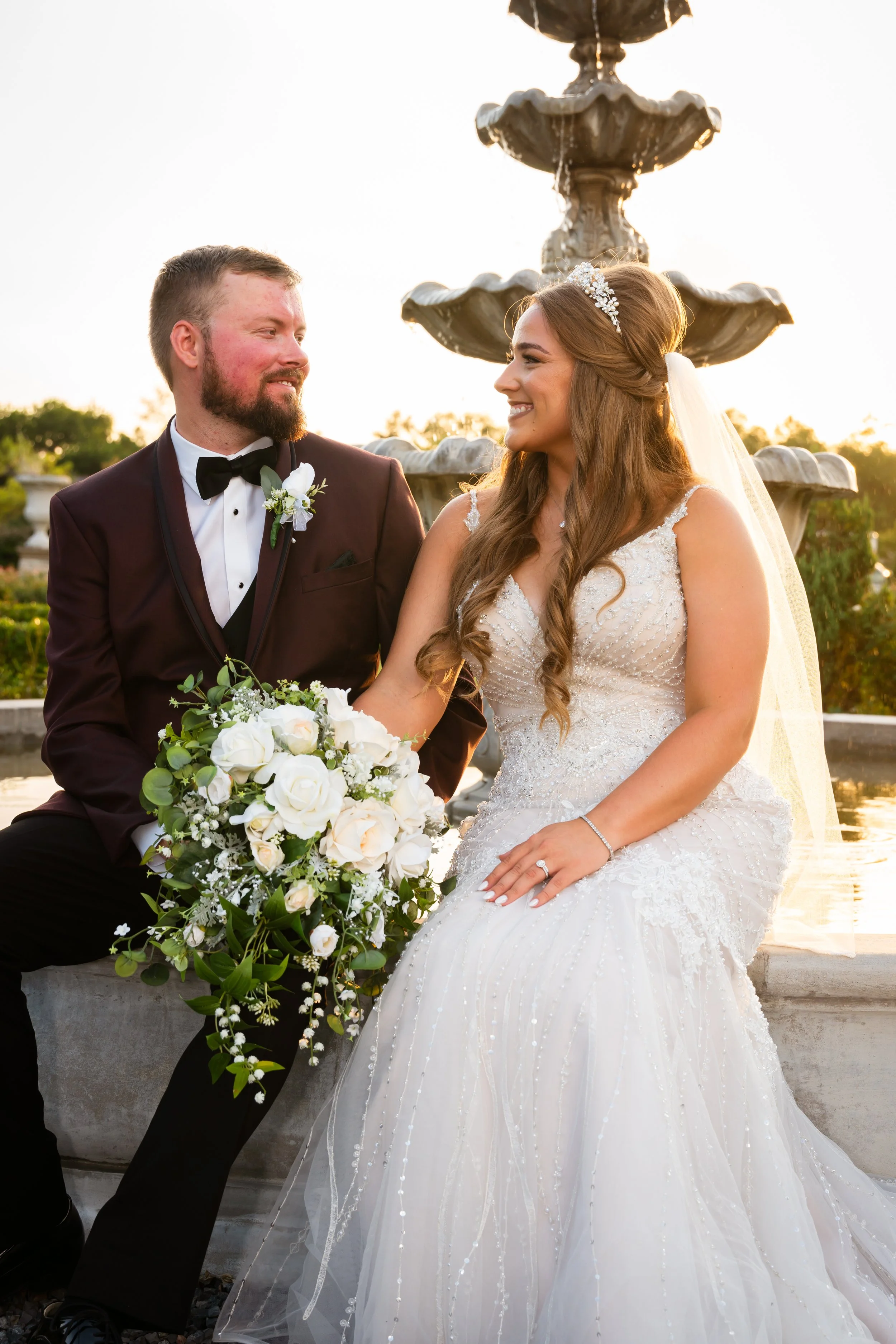 A bride and groom sitting beside each other near a fountain during sunset, smiling and looking at each other. The bride is in a white wedding gown holding a bouquet of white roses, and the groom is in a dark tuxedo with a bow tie.