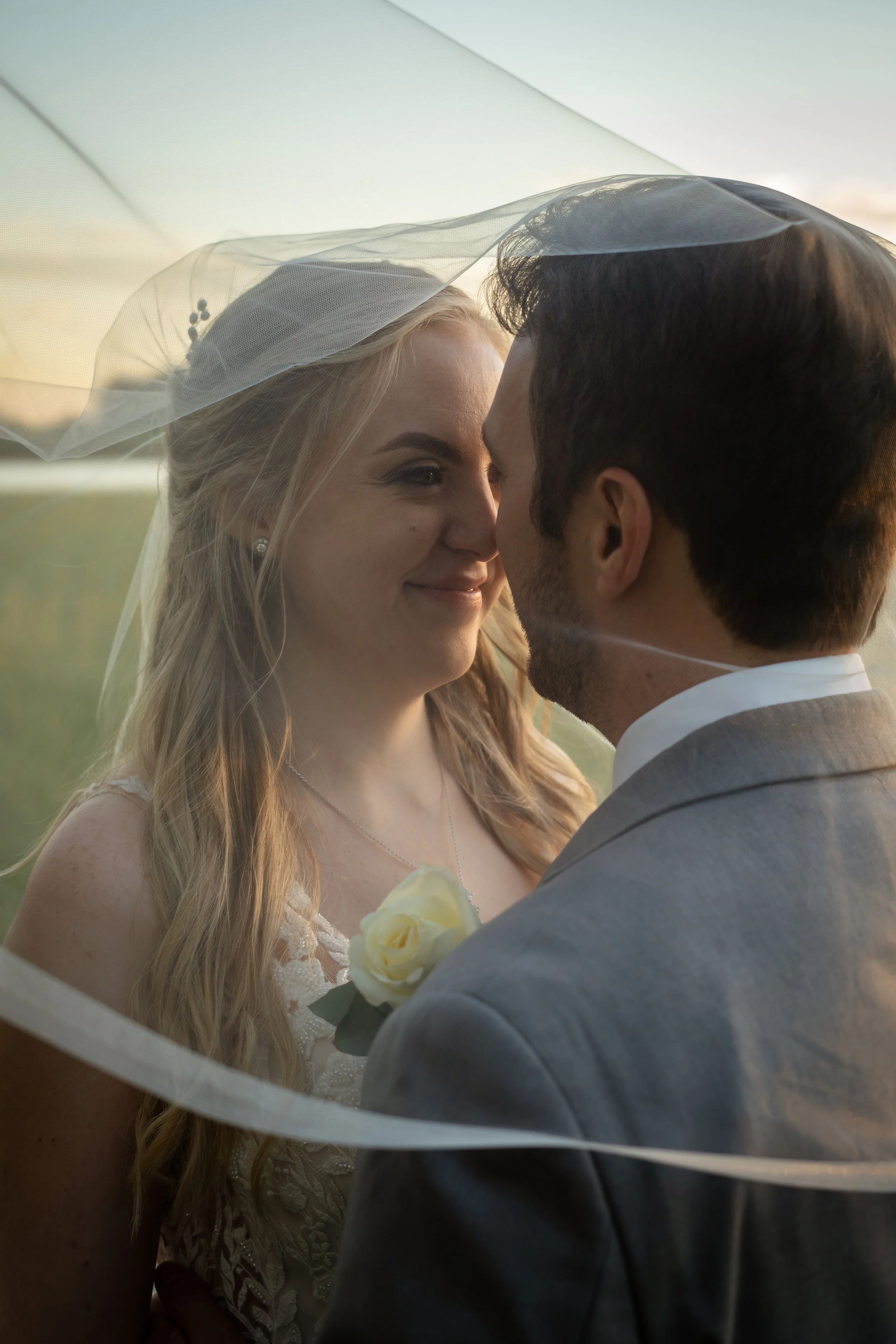 A bride and groom close together outdoors, with the bride wearing a veil and holding a white rose, seen during sunset.