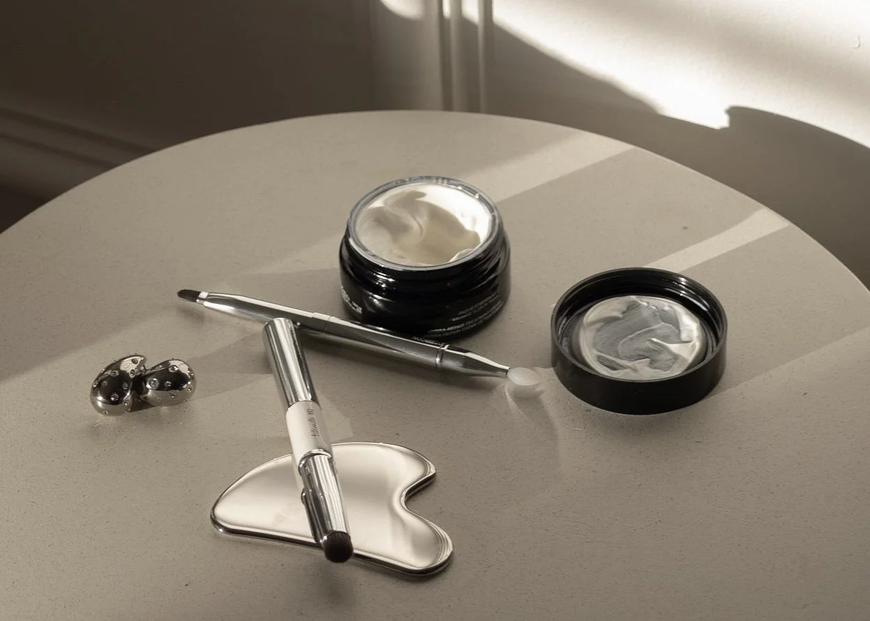 Open jar of white cream, closed jar of similar cream, small metallic massage rollers, metallic spatula, and a white ceramic heart-shaped dish on a light-colored round table, with sunlight casting shadows.