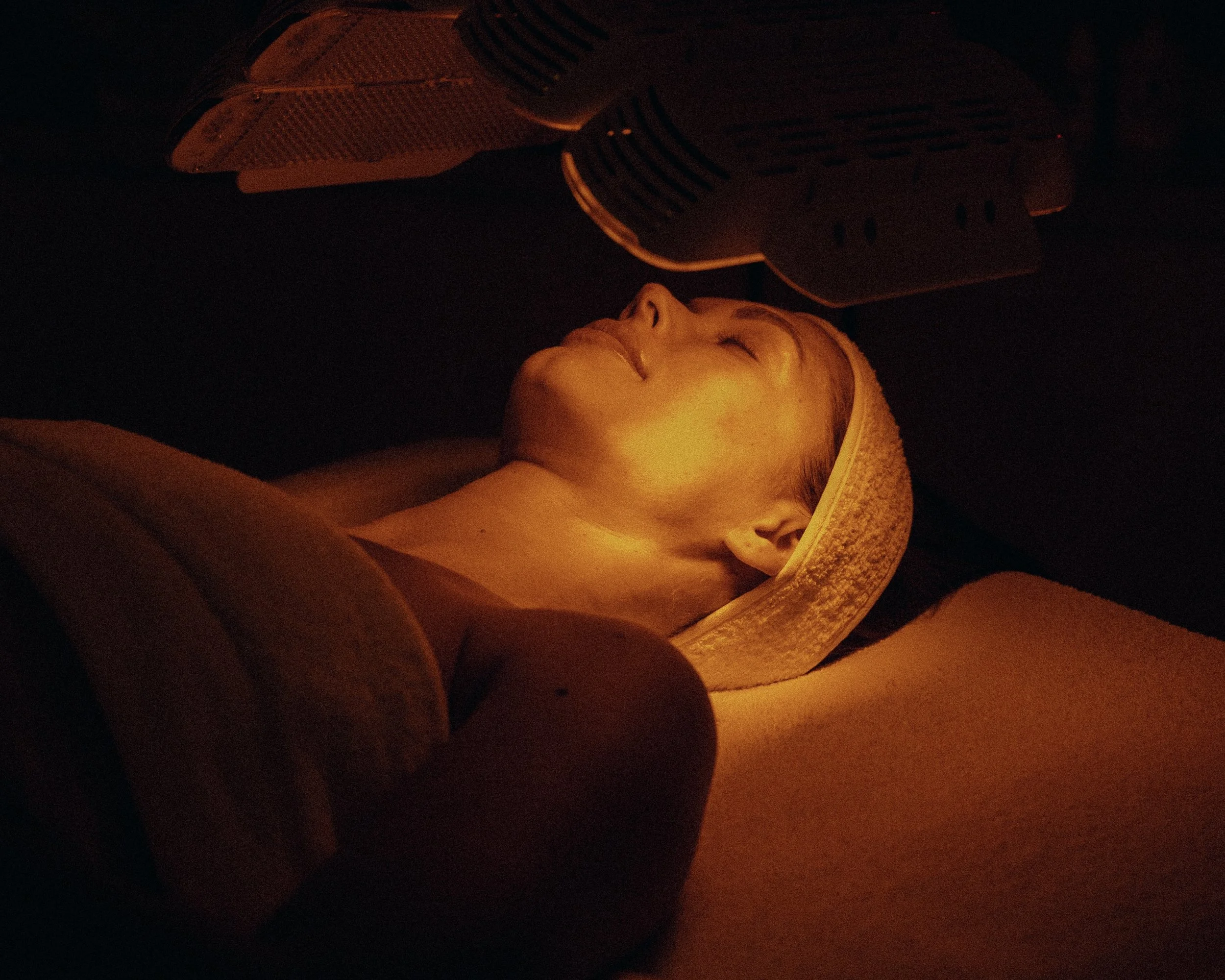 A woman receiving a near-infrared light therapy treatment. She is lying down with her eyes closed, wearing a headband, with a device above her face emitting orange light.