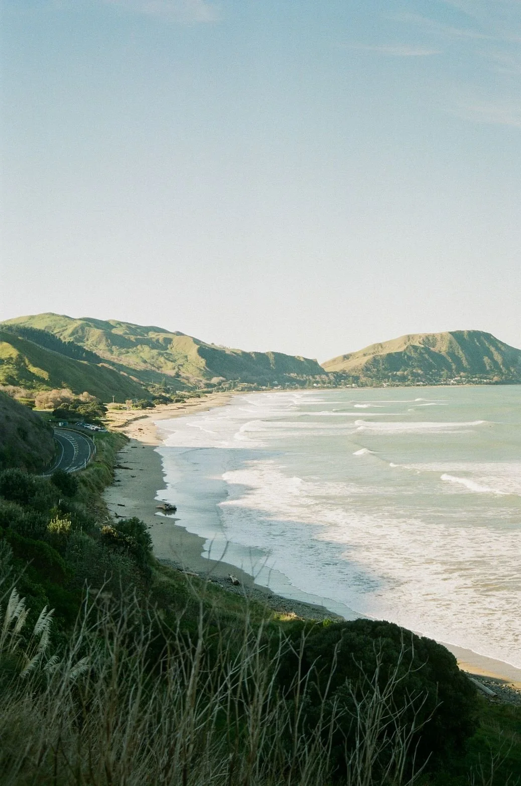A coastal landscape with grassy hills on the left and a beach with ocean waves on the right, along a winding road.