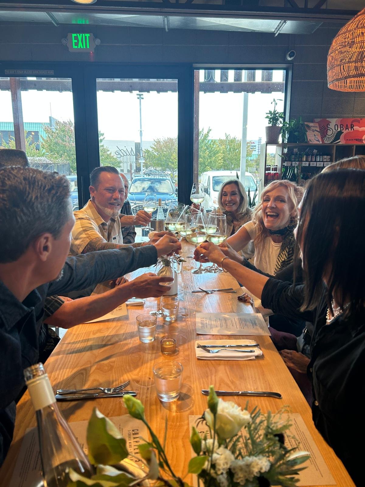 Group of friends raising glasses of wine in a toast at a restaurant table with flowers, menus, and silverware, inside a modern restaurant with large windows and an outdoor view.