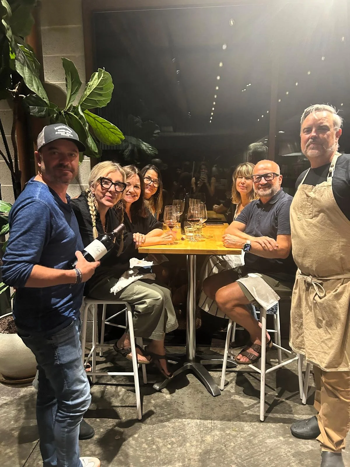 Group of seven people sitting and standing around a high table at a restaurant, with wine glasses on the table and a person holding a bottle of wine. They are indoors with a large plant and dark background.