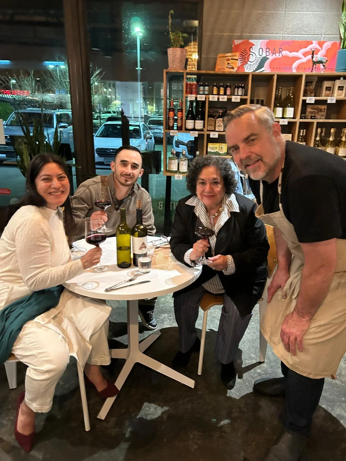Four people sitting around a table in a restaurant, drinking red wine, with a male waiter standing beside the table. The group appears to be enjoying a meal together. Behind them is a large window showing parked cars outside and shelves with bottles 