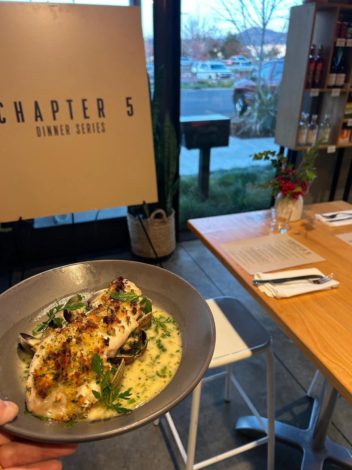 A plate of fish topped with breadcrumbs and herbs, served with a creamy sauce with parsley garnish, held inside a restaurant near a window overlooking parked cars and trees.