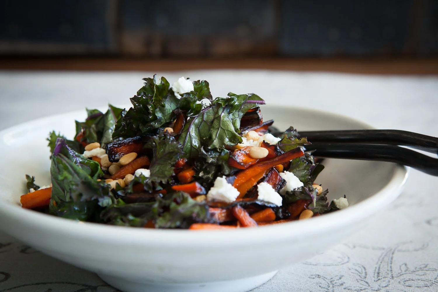 A bowl of mixed vegetable salad with leafy greens, shredded carrots, pine nuts, and crumbled cheese, with black chopsticks resting on the side.