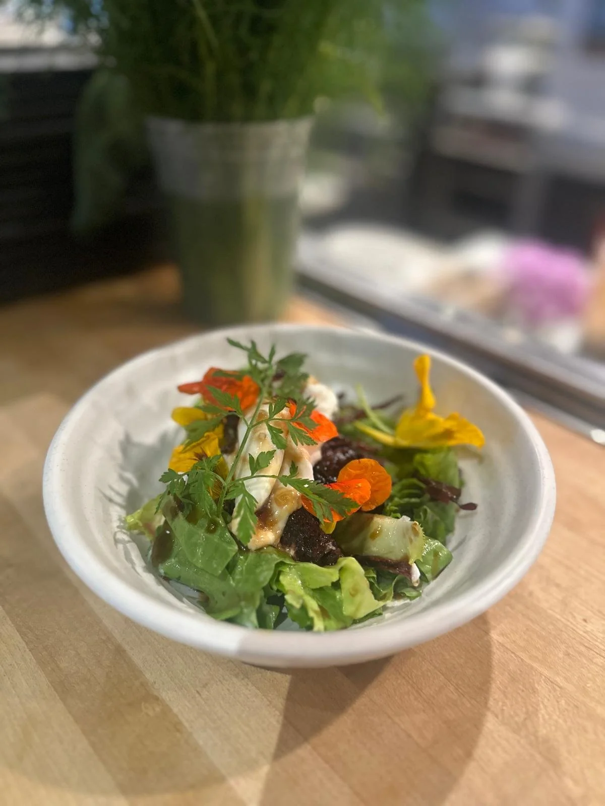 Fresh mixed green salad with edible flowers, topped with herbs, drizzled with dressing in a white bowl, with a vase of green foliage in the background on a wooden surface.