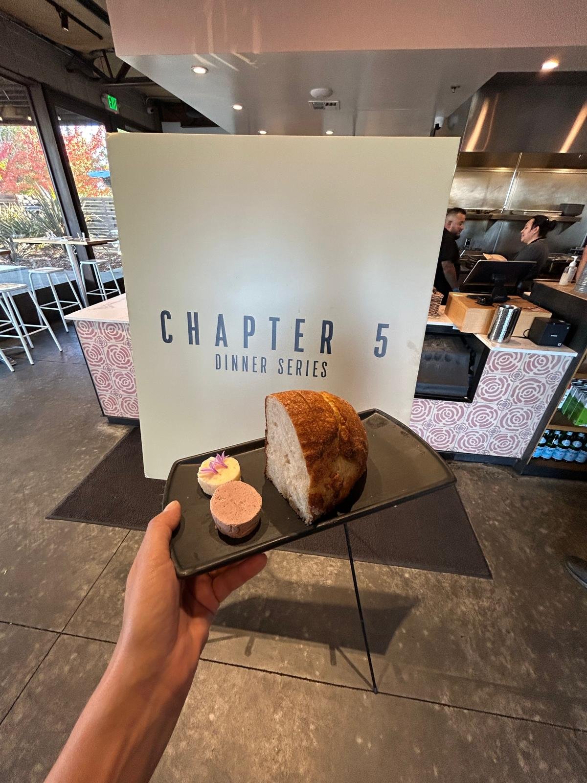 A person holding a black plate with a large slice of cake and two small desserts, in front of a sign that says 'Chapter 5 Dinner Series' inside a restaurant.