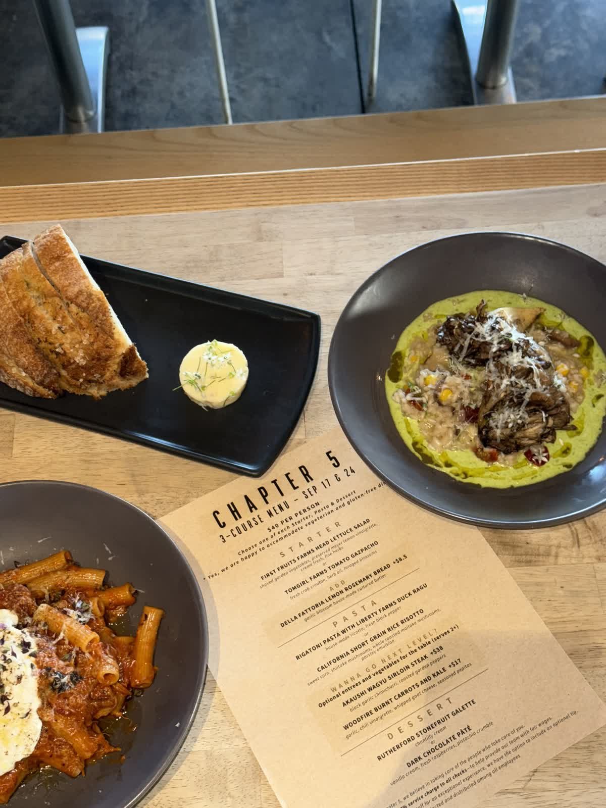 A table set with three dishes: a black plate with toasted bread and a small pat of butter, a black bowl with a green sauce or puree topped with meat and grated cheese, and a gray bowl with pasta and sauce. There is also a restaurant menu on the table