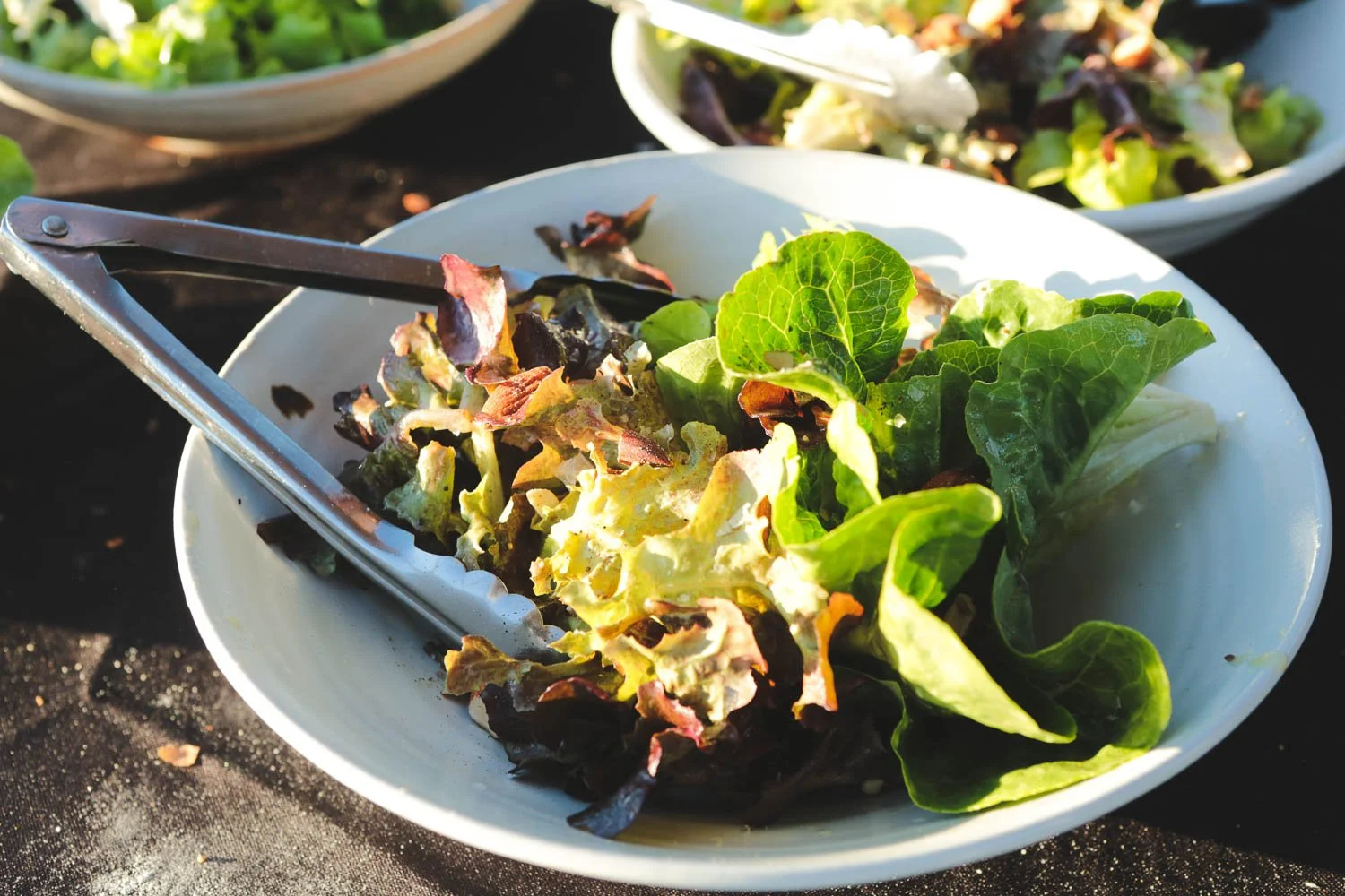 Close-up of a white bowl filled with mixed salad greens, including lettuce and other leafy greens, with a pair of metal tongs resting on the edge, on a black surface with sunlight shining