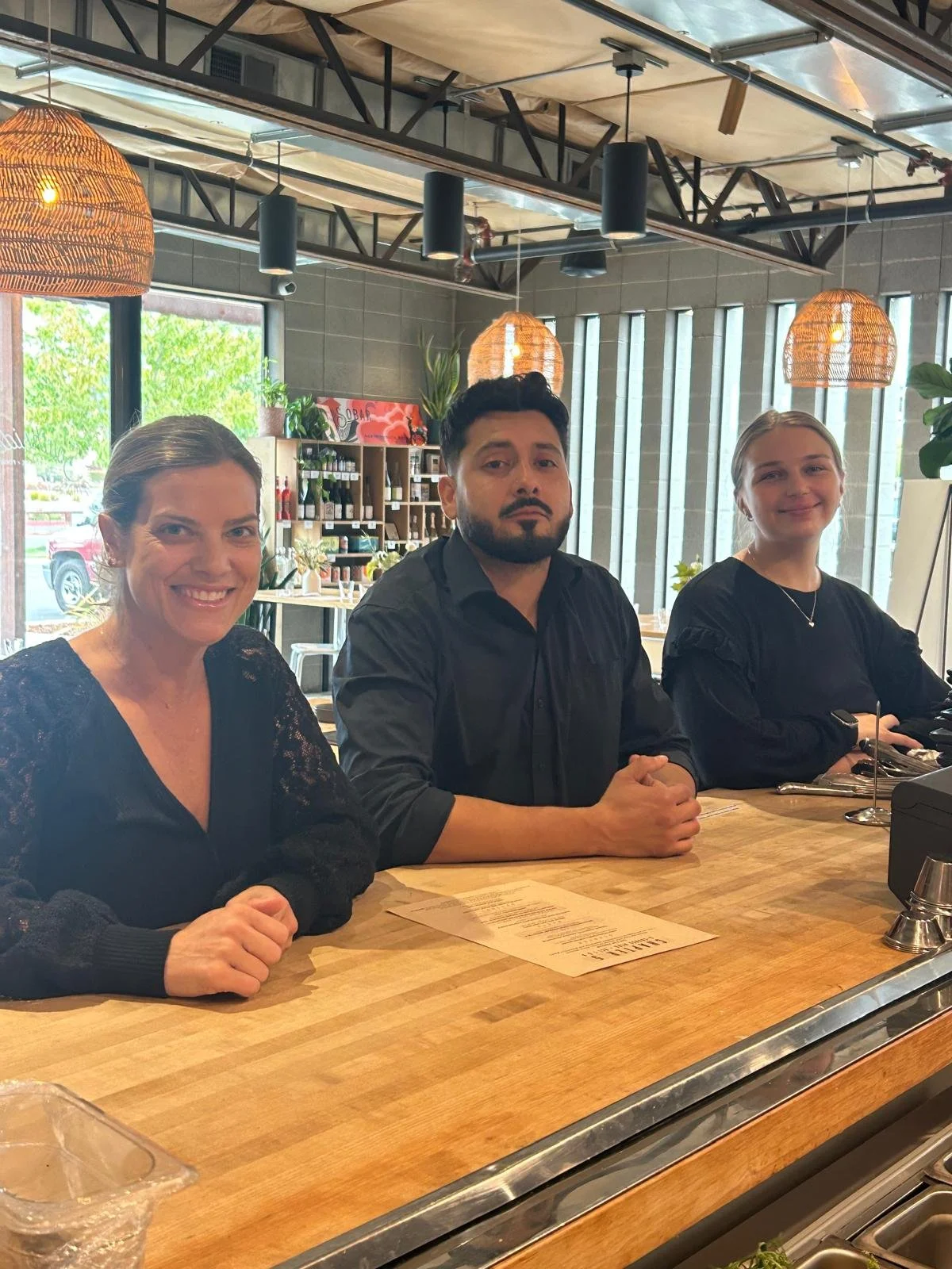 Three people sitting at a restaurant or café table with menus and utensils, inside a modern space with large windows, hanging lights, and decorative plants.